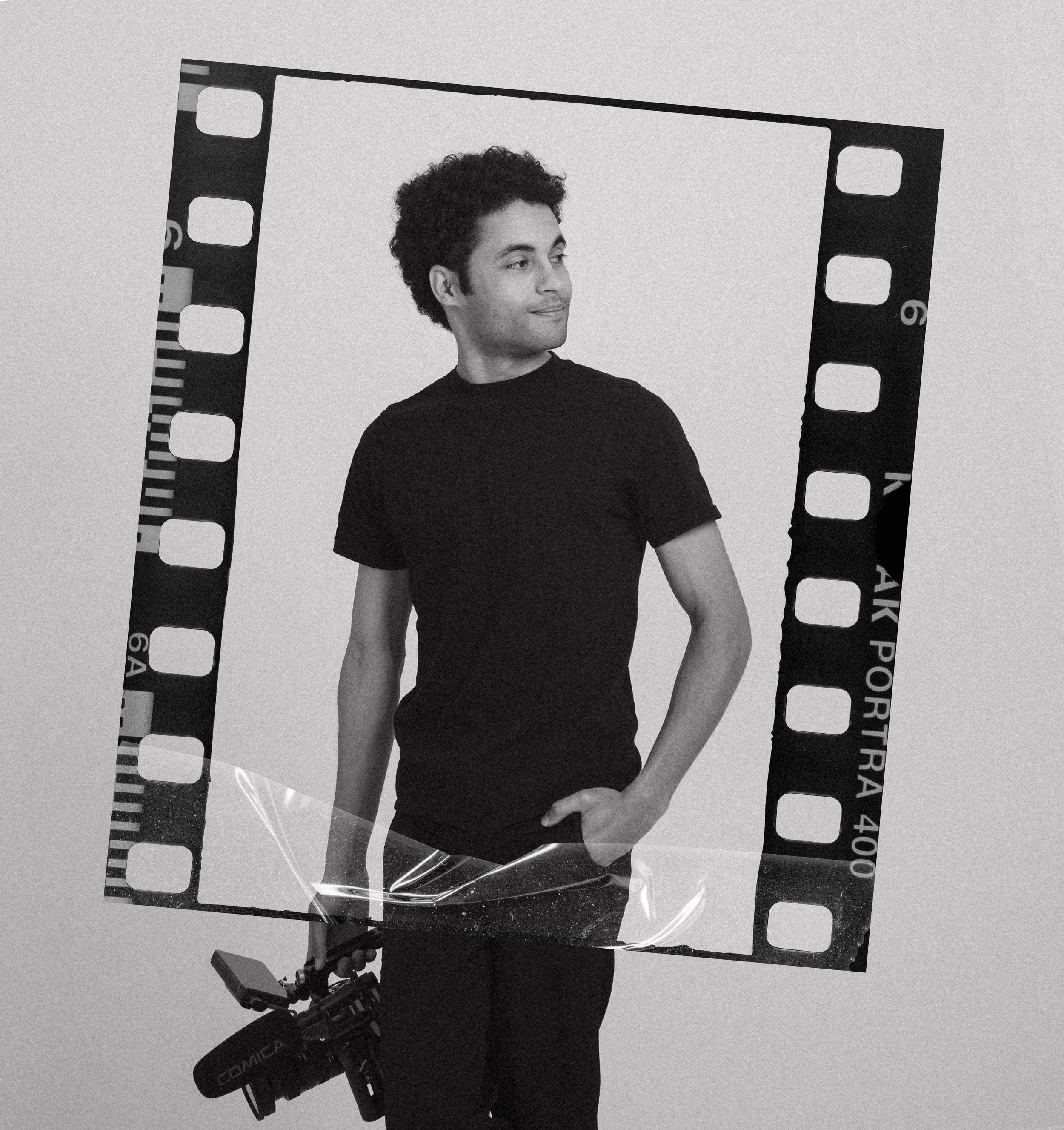 Black and white photo of a young man with curly hair wearing a black t-shirt, standing inside a large film strip frame, looking to his right with a slight smile, against a plain light-colored background.