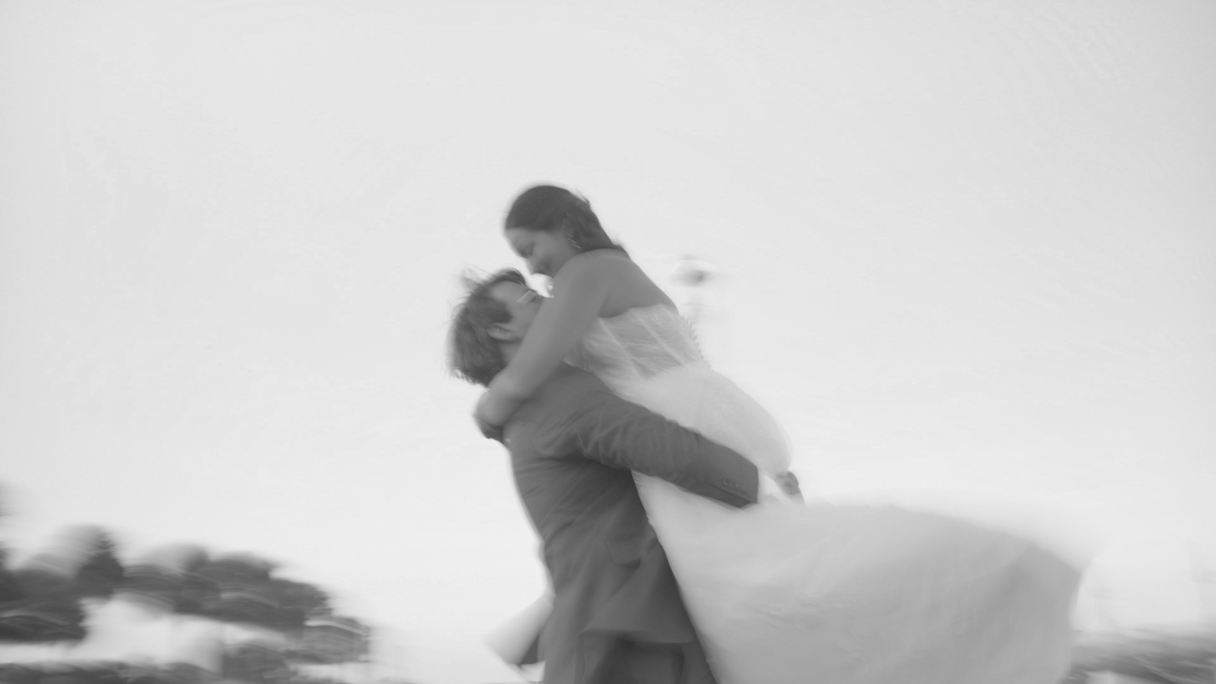 A couple in wedding attire, with the groom lifting the bride off the ground in a joyful embrace outdoors during daytime.
