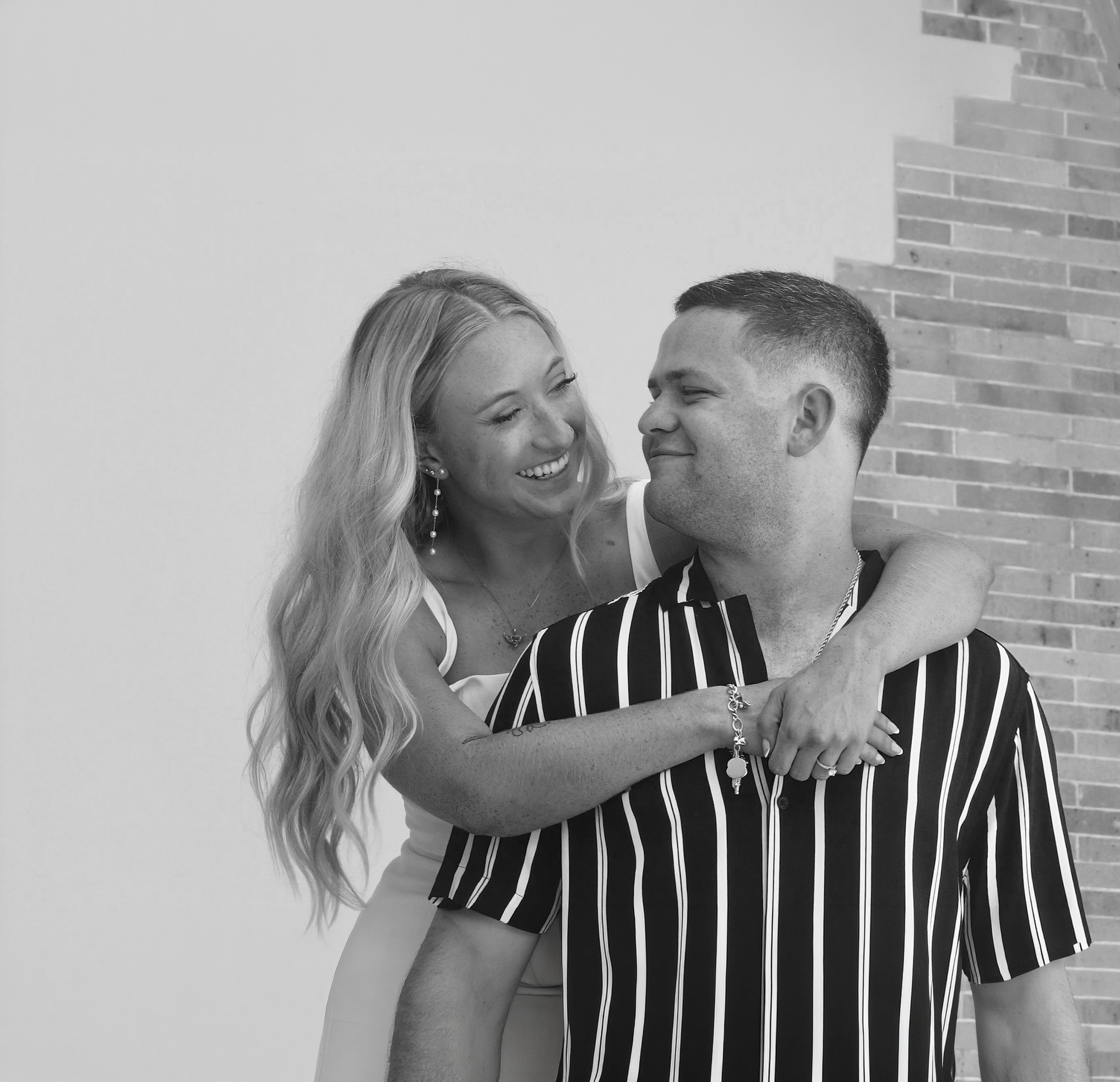 Black and white photo of a smiling woman with long wavy hair embracing a man with short hair and a striped shirt, against a background of a brick wall.