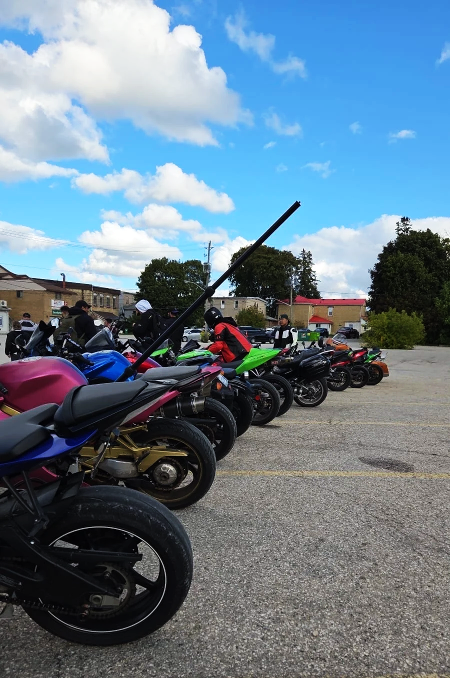 Multiple motorcycles parked in a lot with people wearing riding gear, and a blue sky with clouds overhead.