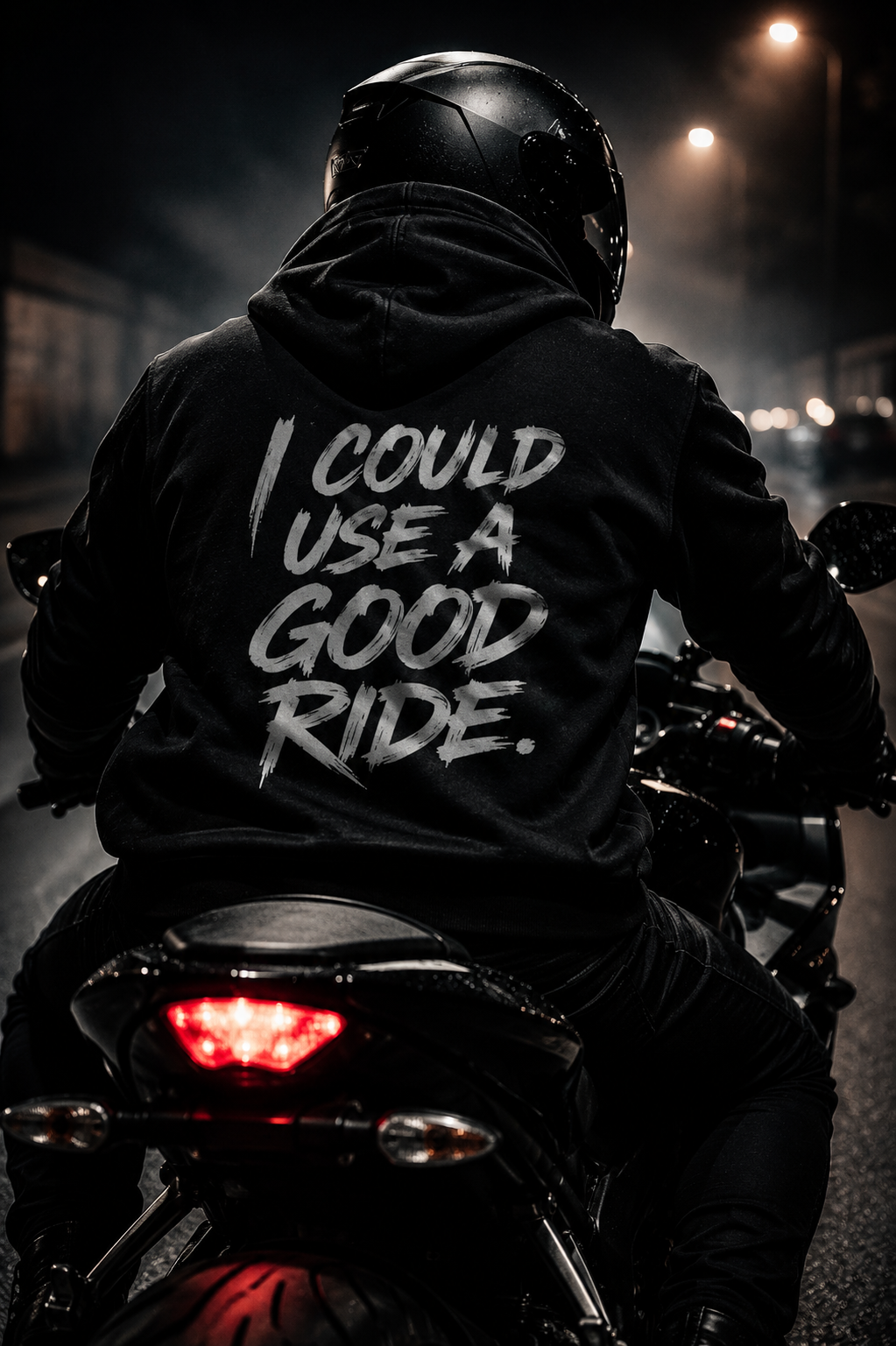 Motorcycle rider wearing a black helmet and hoodie, riding on a dark city street at night, with the back of the hoodie displaying the words 'I COULD USE A GOOD RIDE' in bold, distressed font.