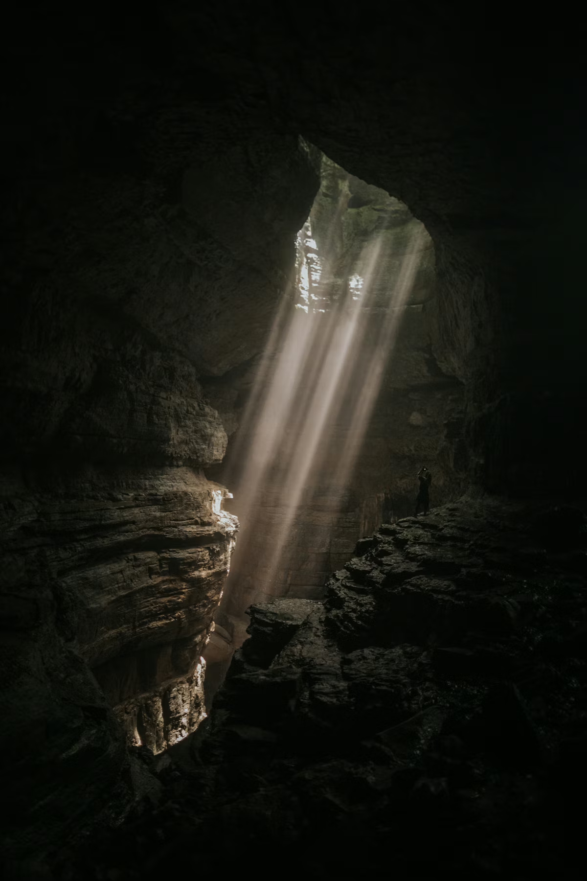 Sunlight streams into a dark, rocky canyon creating beams of light illuminating the interior, with a person standing on the right side.