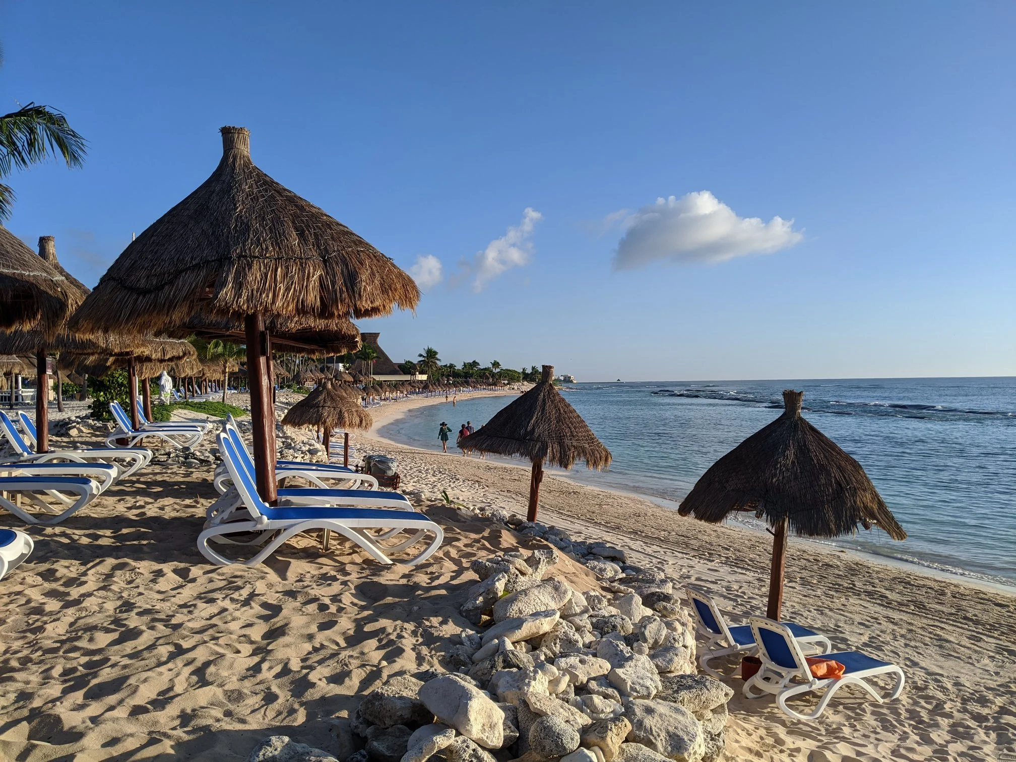 A beach with straw umbrellas and white lounge chairs on sandy shore, ocean waves and blue sky with a few clouds in the background.