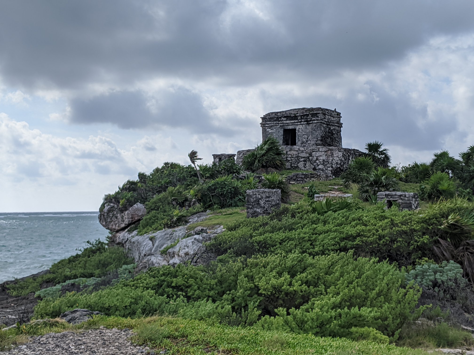 Ancient stone structure on a hillside surrounded by green bushes and trees, cloudy sky overhead.