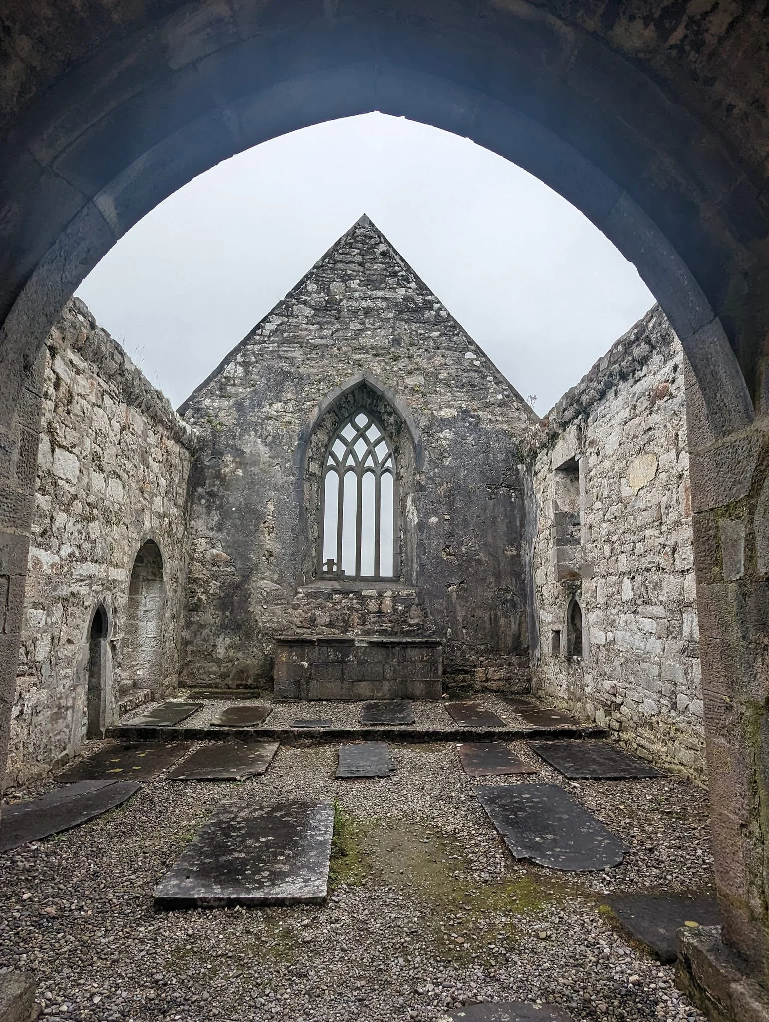 The interior of an old stone church or chapel with standing water on the floor. Tall walls with arched windows and a peaked roof visible through a large archway