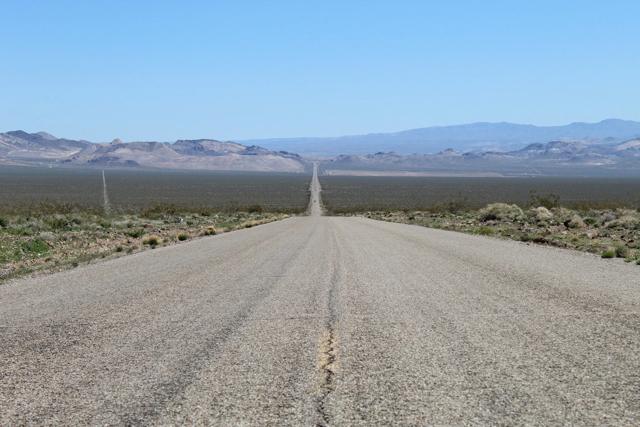 A long, straight road stretching into the desert with mountains in the background under a clear blue sky.