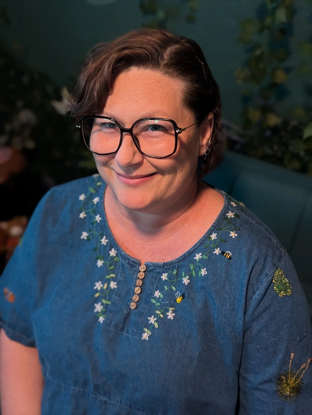 A woman with short brown hair and glasses, smiling, wearing a blue embroidered shirt with flowers and bees, in front of a dark background with green foliage.