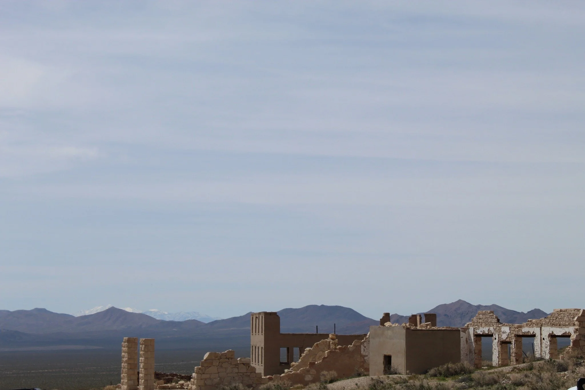 Ruins of an ancient stone building in a desert landscape with mountains and snow-capped peaks in the distance under a partly cloudy sky.