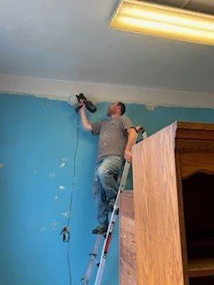 A man standing on a ladder painting the upper wall of a room with light blue paint, using a roller brush.