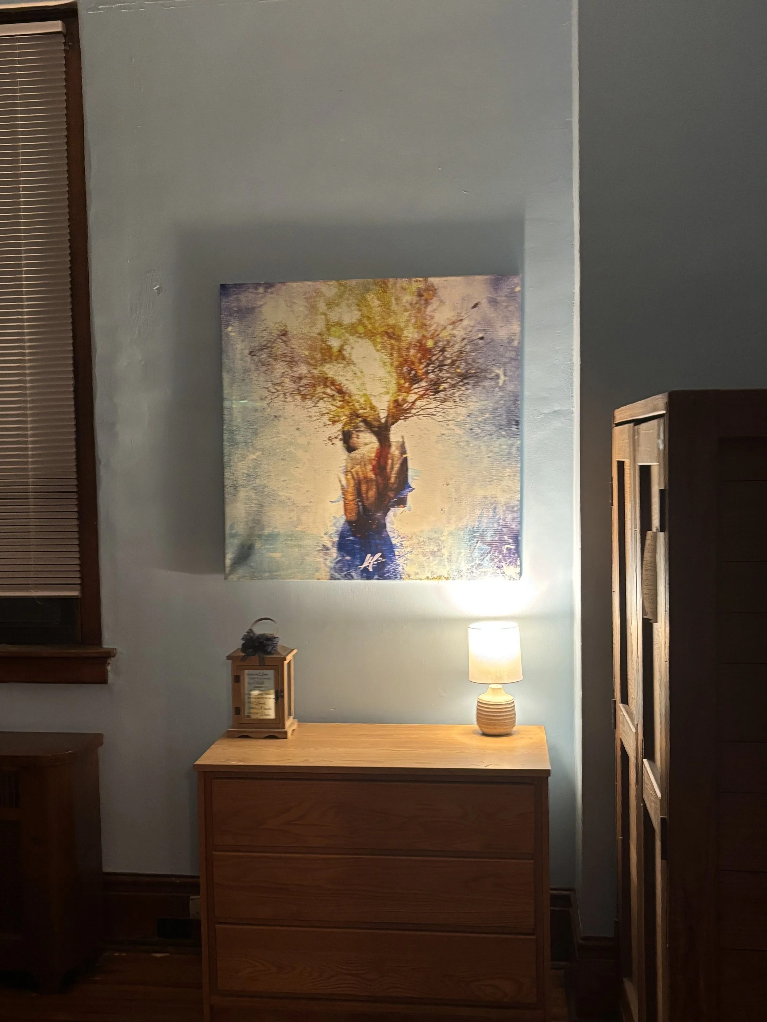 A framed artwork of a person standing under a tree with colorful leaves, hanging on a light blue wall. Below the artwork, there is a wooden dresser with a small lantern and a table lamp turned on, emitting warm light.