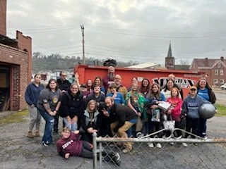 Group of people posing outdoors on a cloudy day, standing in front of a red truck with a small vintage train car and locomotive. Some individuals are holding helmets and standing near a bicycle, with a church steeple visible in the background.