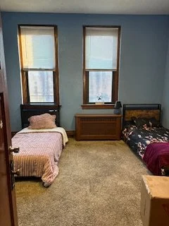 A bedroom with two windows, two beds with bedding, and a radiator between the windows.