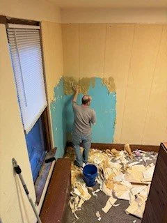 Person painting a wall with blue paint in a room with beige walls, window, and peeling wallpaper on the floor.