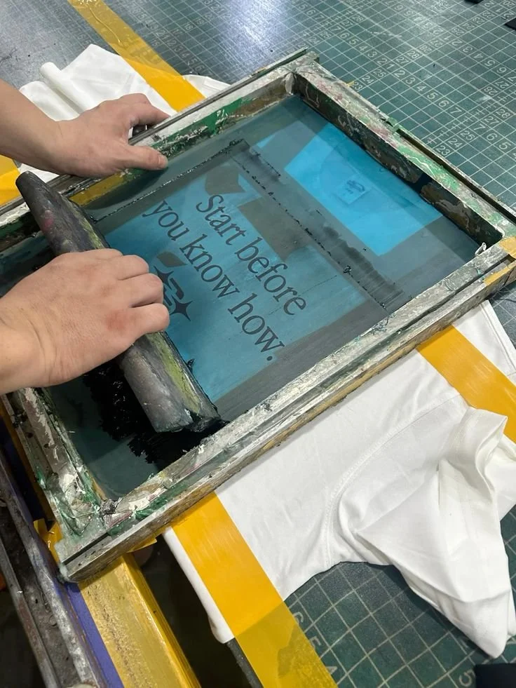 A person is screen printing a design onto a white T-shirt. The design on the screen reads 'Start before you know how 25'. The setup includes a printing frame, a squeegee, and a worktable with a green gridded cutting mat.