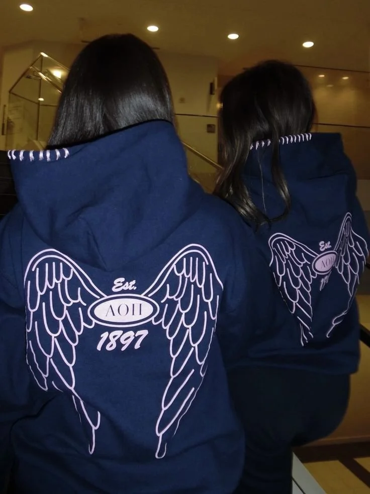 Two people wearing navy blue hoodies with angel wings and Greek letters on the back. The setting appears to be indoors with warm lighting.