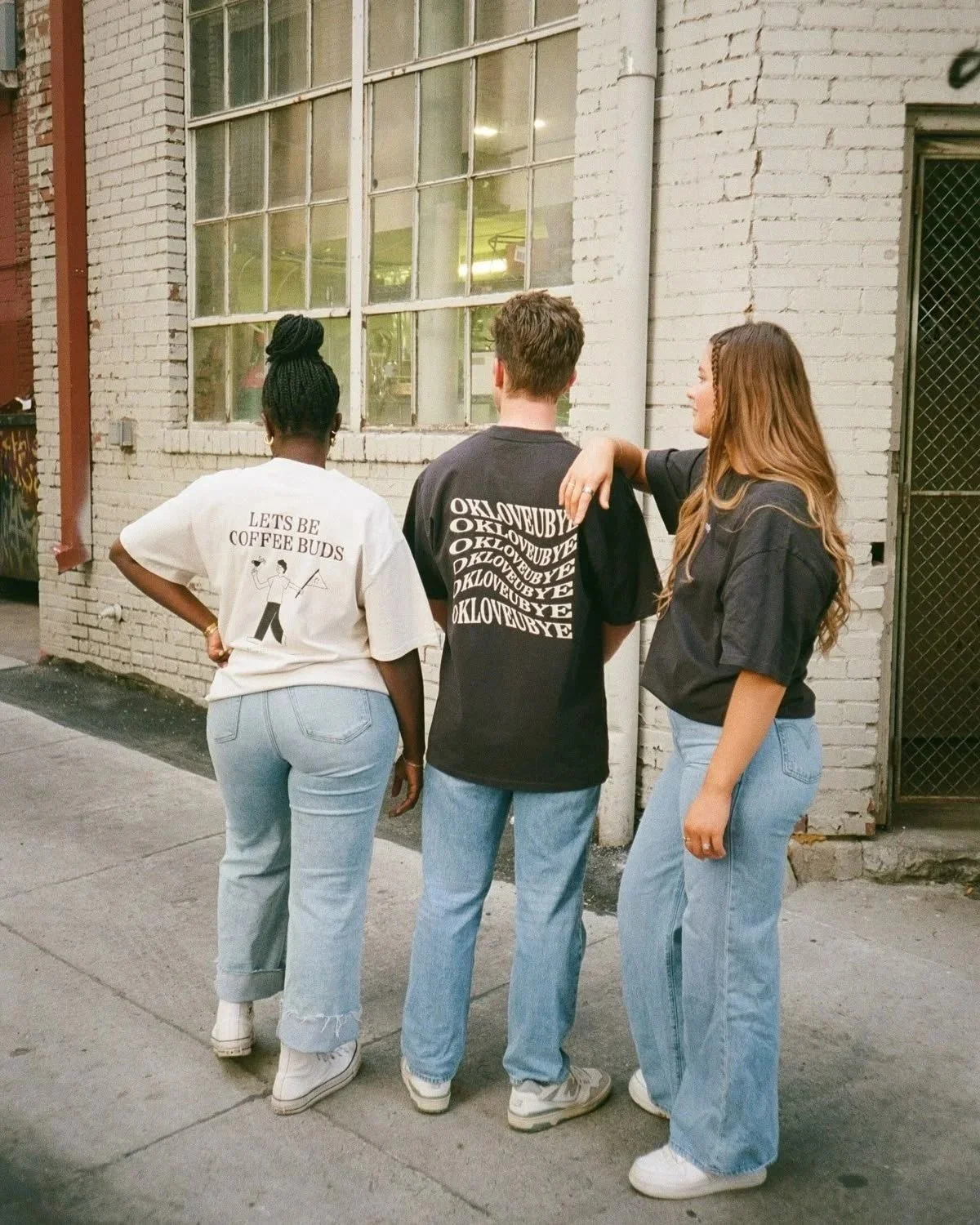 Three people standing on a sidewalk outside a brick building with a large window, engaging in conversation.