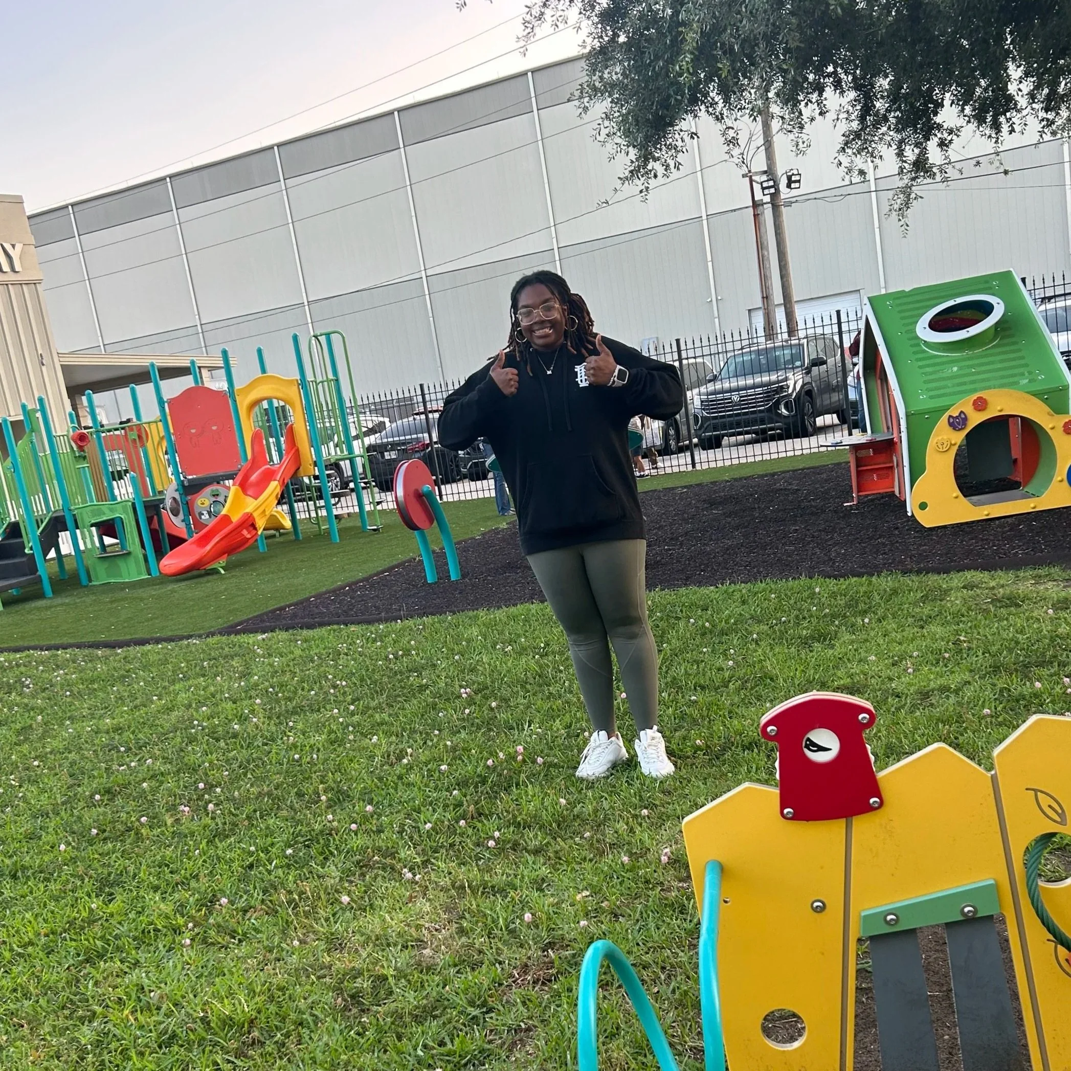 A woman smiling and making peace signs in a playground with colorful play structures, grass, and a fence.