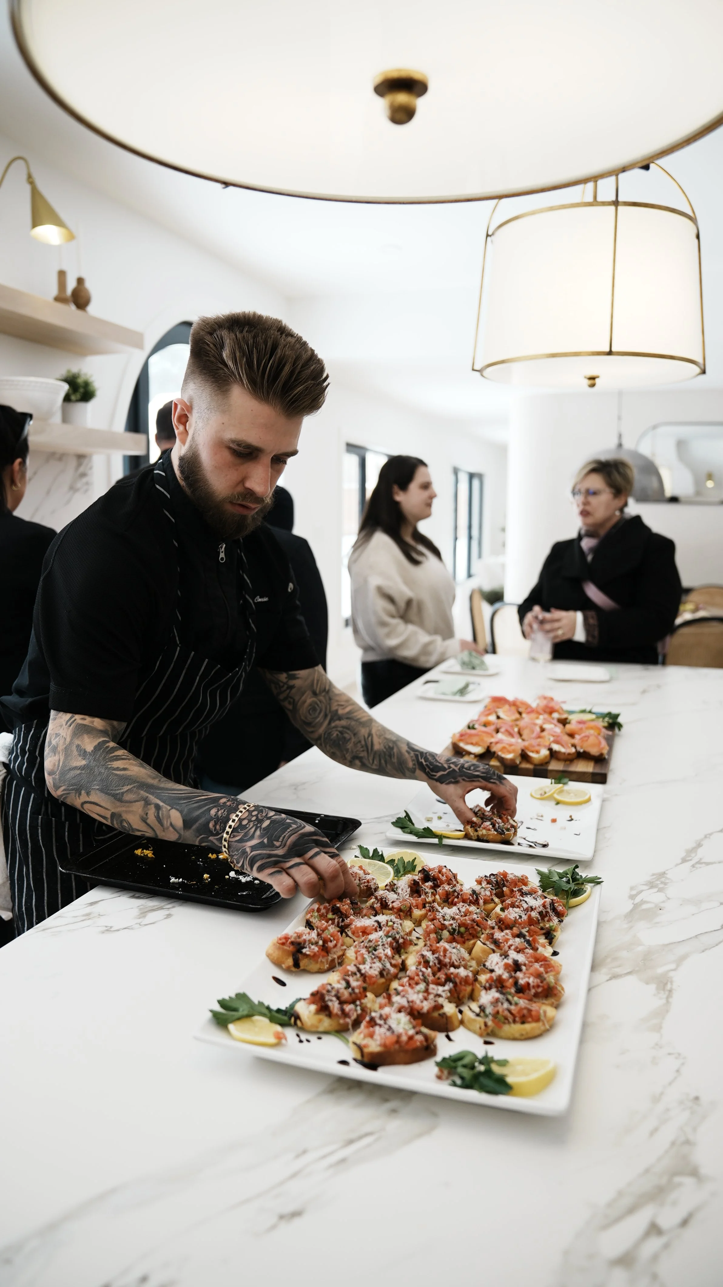 A tattooed man in a black apron preparing and serving thin-crust pizza slices topped with tomatoes, herbs, and cheese on a white marble countertop as people chat in the background.