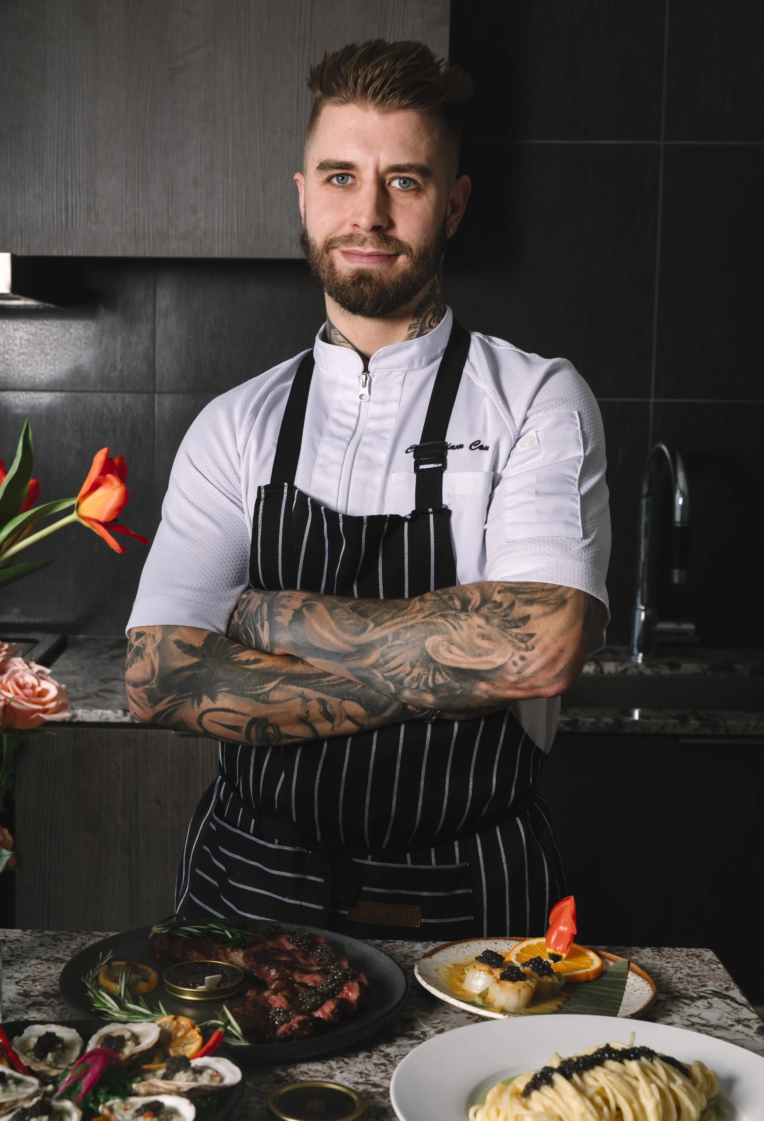 A male chef with tattoos standing with arms crossed in a modern kitchen, surrounded by plates of gourmet food.