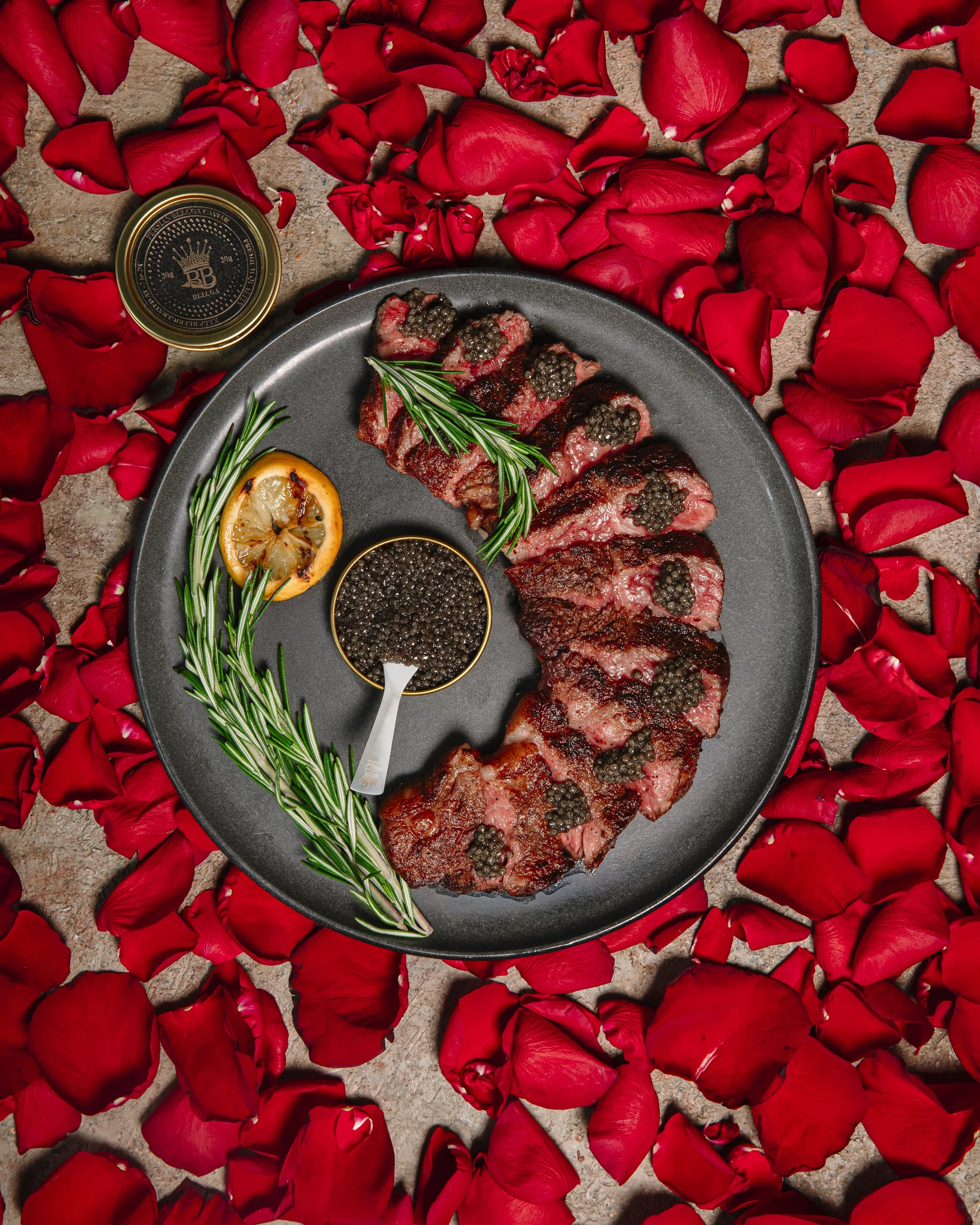 Seared steak slices topped with black caviar on a black plate, garnished with a sprig of rosemary, lemon slice, and a small bowl of black caviar, surrounded by red rose petals.