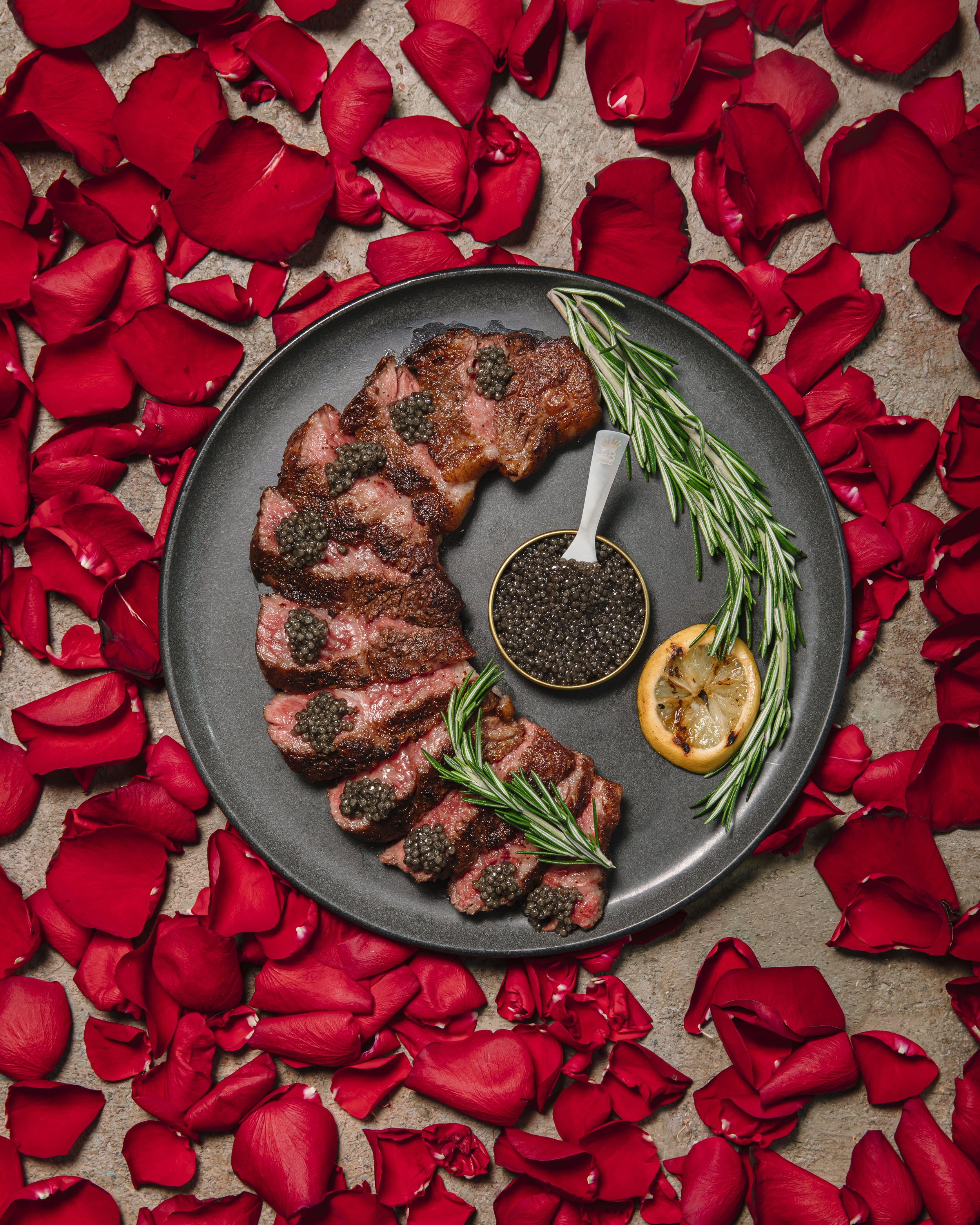 Sliced steak topped with black peppercorns on a black plate, garnished with sprigs of rosemary and lemon slices, surrounded by red rose petals.