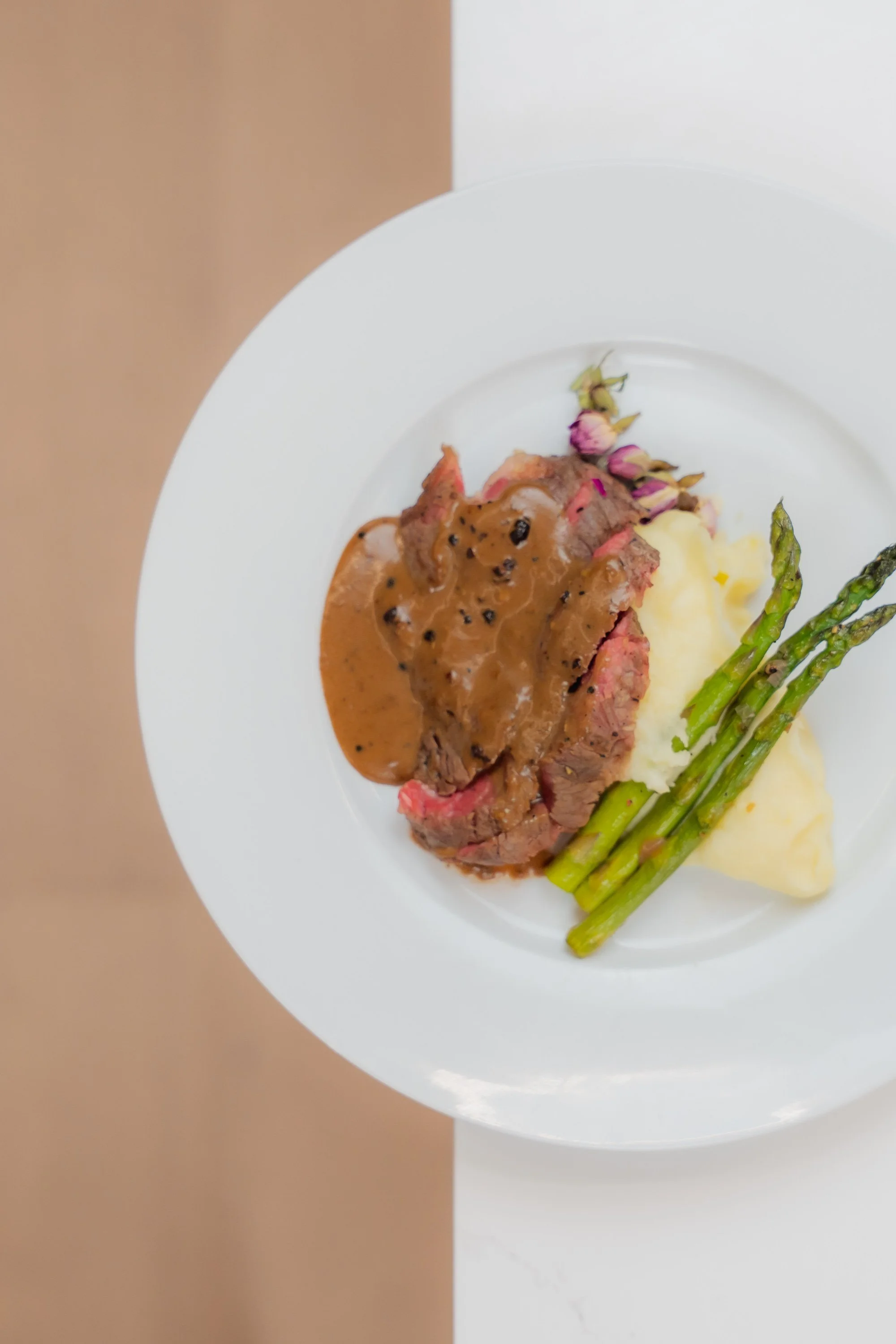 A white plate with slices of beef topped with gravy, alongside asparagus spears, mashed potatoes, and a few pink and green flower petals.