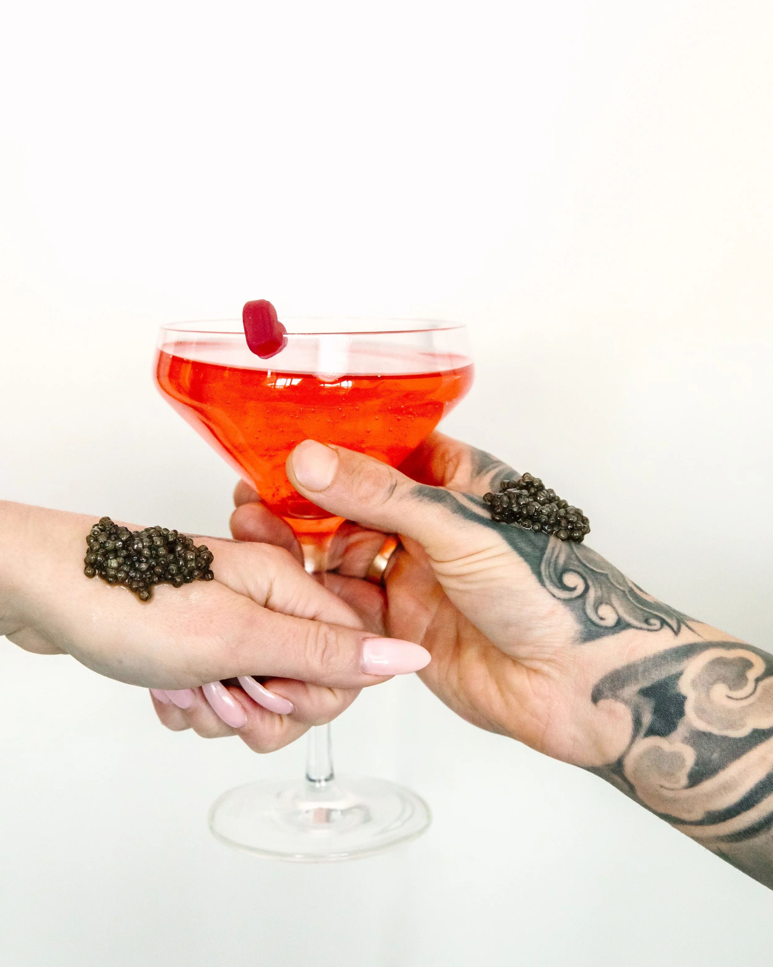 Two tattooed hands hold a glass of red cocktail with a cherry garnish.