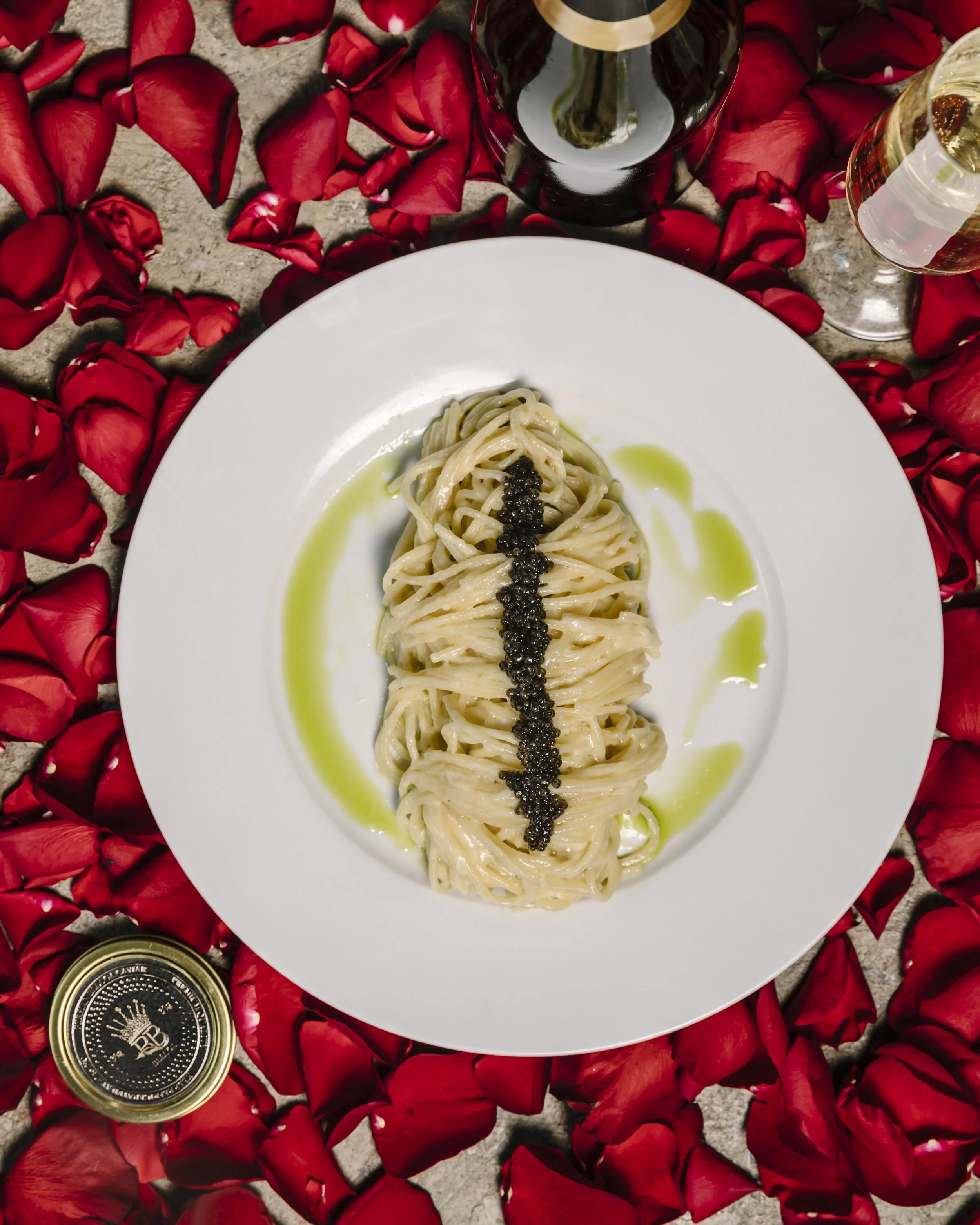 Plate of pasta with black caviar and olive oil, surrounded by red rose petals, with wine glasses and a bottle on a textured surface.