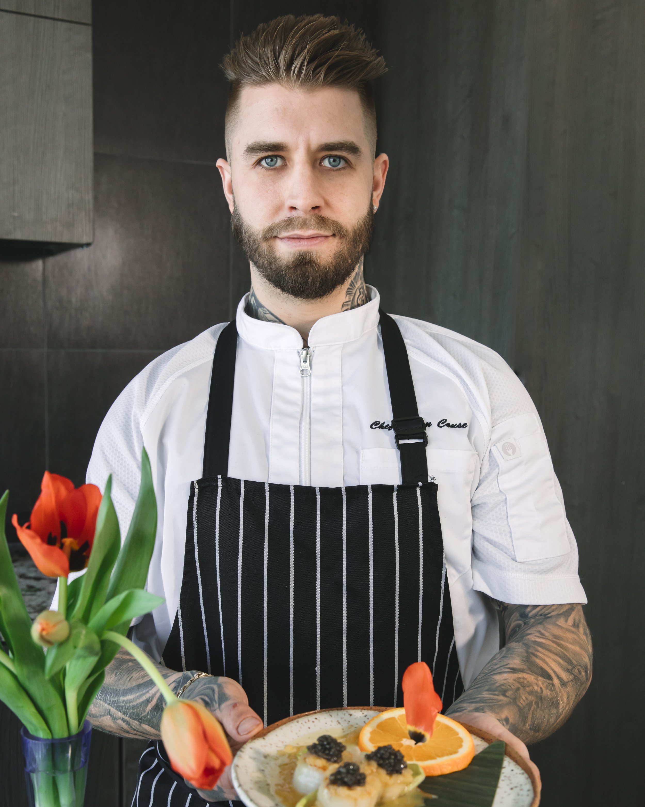 A man with blue eyes, a beard, and tattoos on his arms and neck, dressed in a white chef's coat and black apron, holding a plate of food, with orange tulips on the left side of the image.