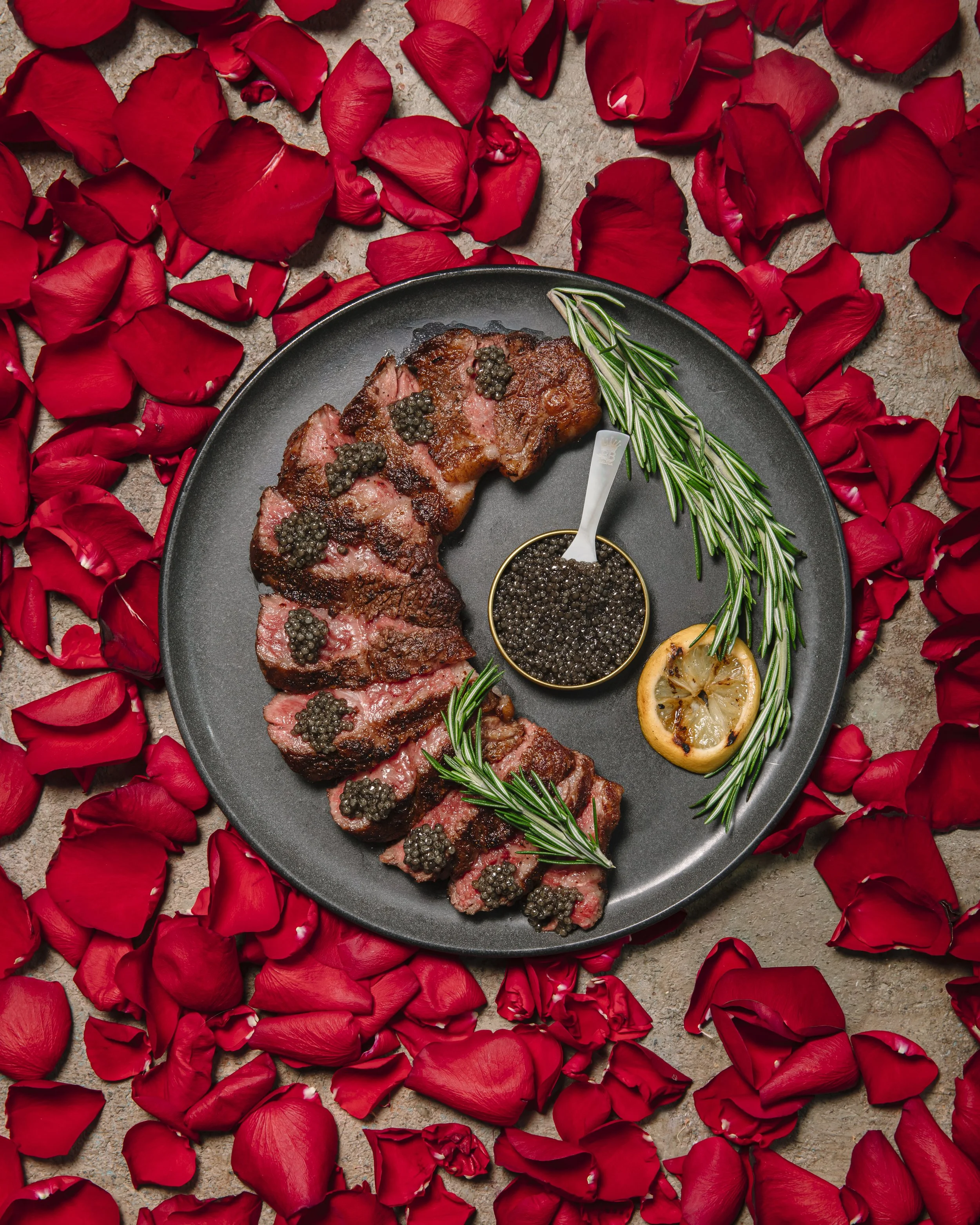 Sliced steak topped with black caviar on a gray plate, garnished with rosemary sprigs, a lemon slice, and a bowl of black caviar, surrounded by red rose petals.