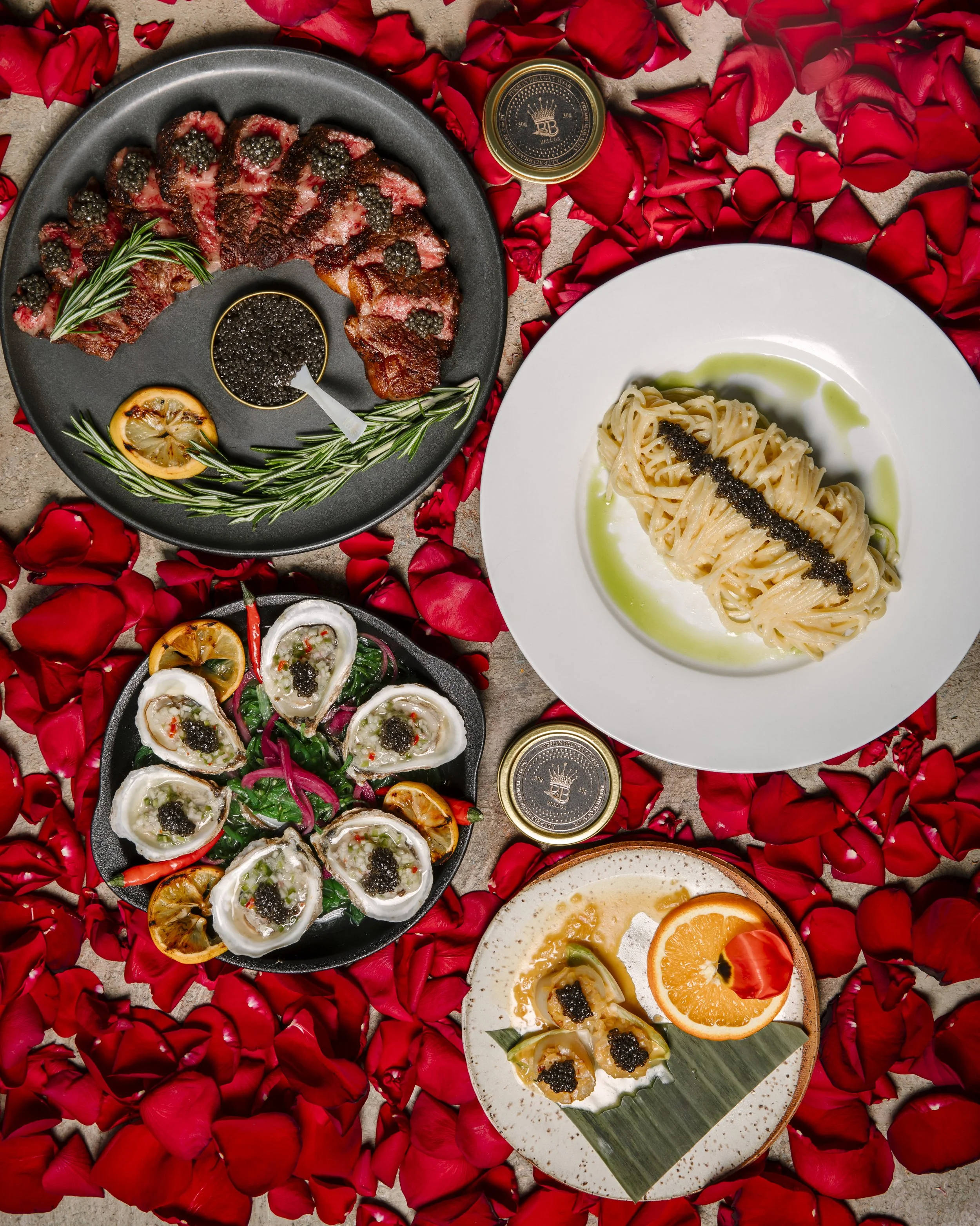 A variety of dishes on a bed of red rose petals, including sliced steak with blackberries, ravioli with green sauce, oysters with garnishes, and a dessert with oranges and whipped cream.