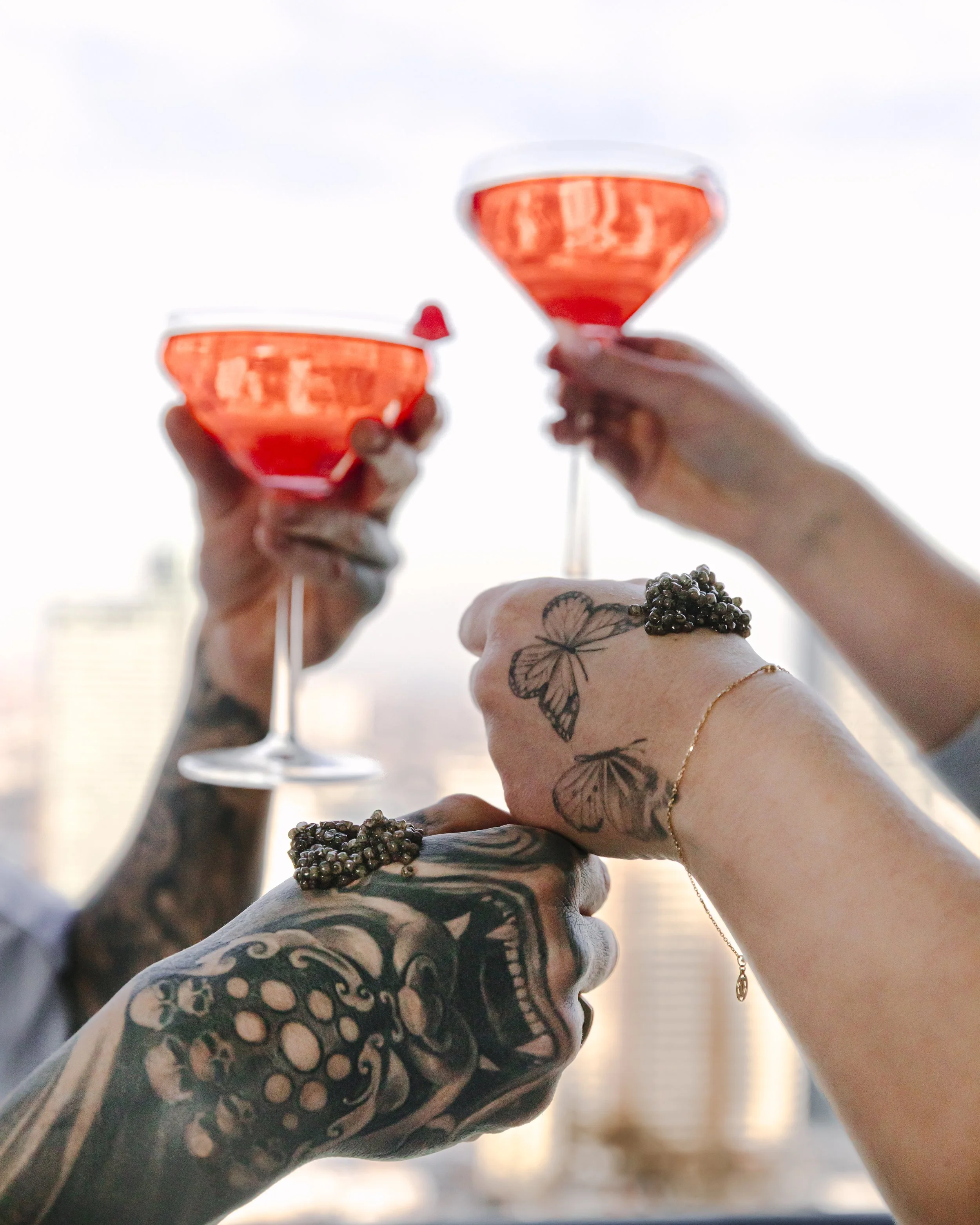 Two people with tattoos holding glasses of pink cocktails, toasting against a cityscape background.
