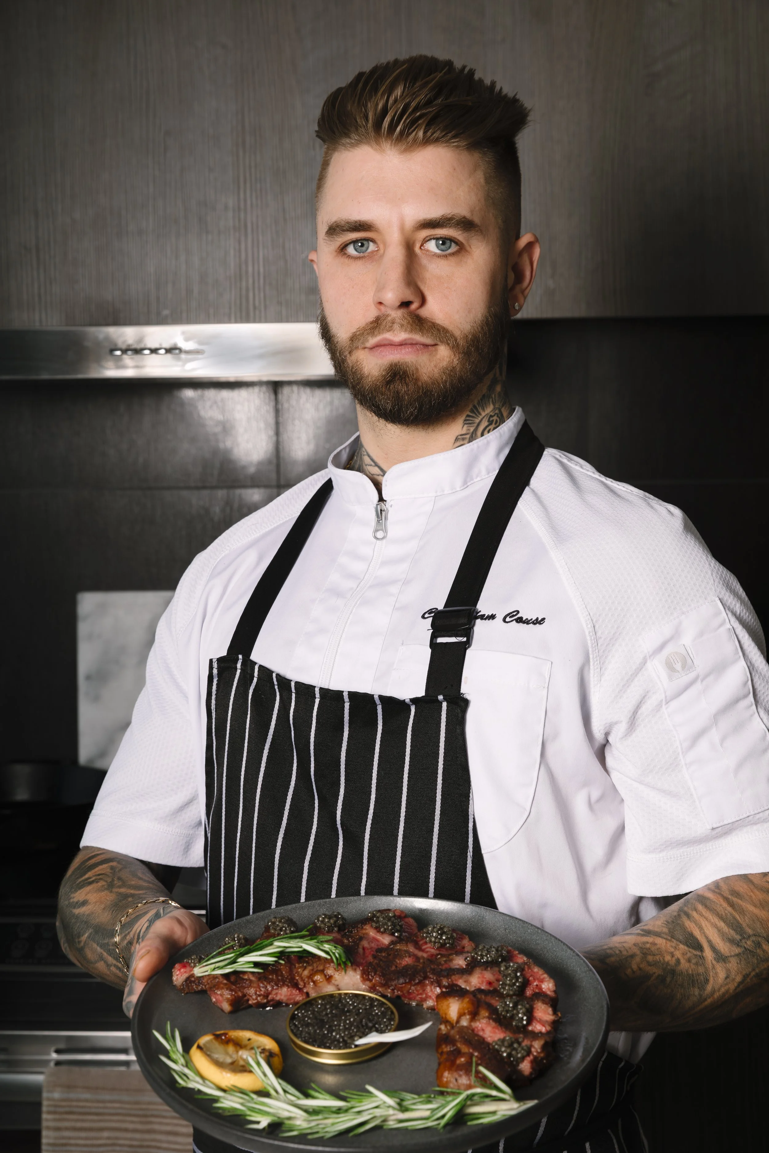 A male chef holding a black plate with cooked steak topped with black caviar, garnished with rosemary sprigs and lemon slices, in a professional kitchen setting.