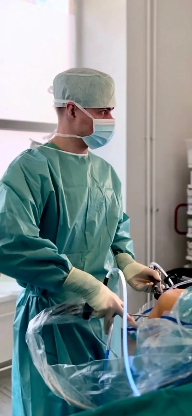 A healthcare professional in surgical attire, including a green gown, cap, mask, and gloves, performing a medical procedure in a clinical setting.