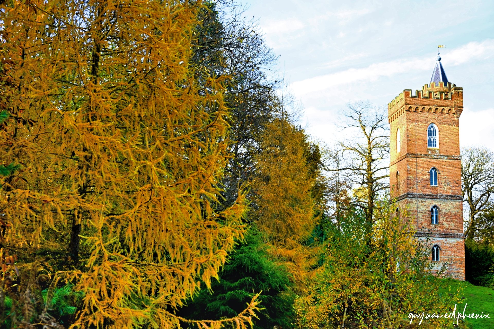 A tall brick tower with arched windows and a decorative top, surrounded by trees with fall foliage in yellow, orange, and green, under a partly cloudy sky.