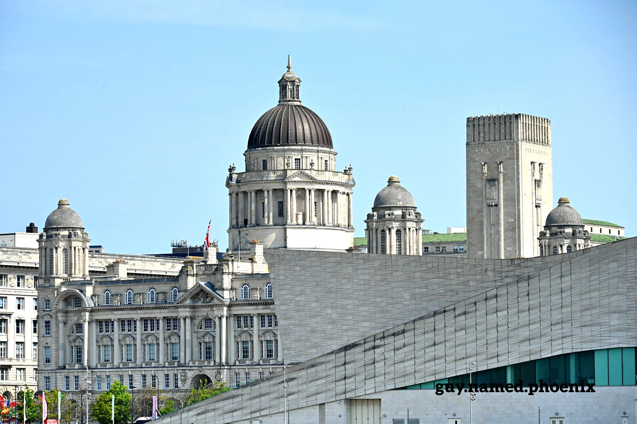 Skyline of historic Port of Liverpool building and modern buildings in Liverpool, England, featuring prominent domes and a tower against a clear blue sky.