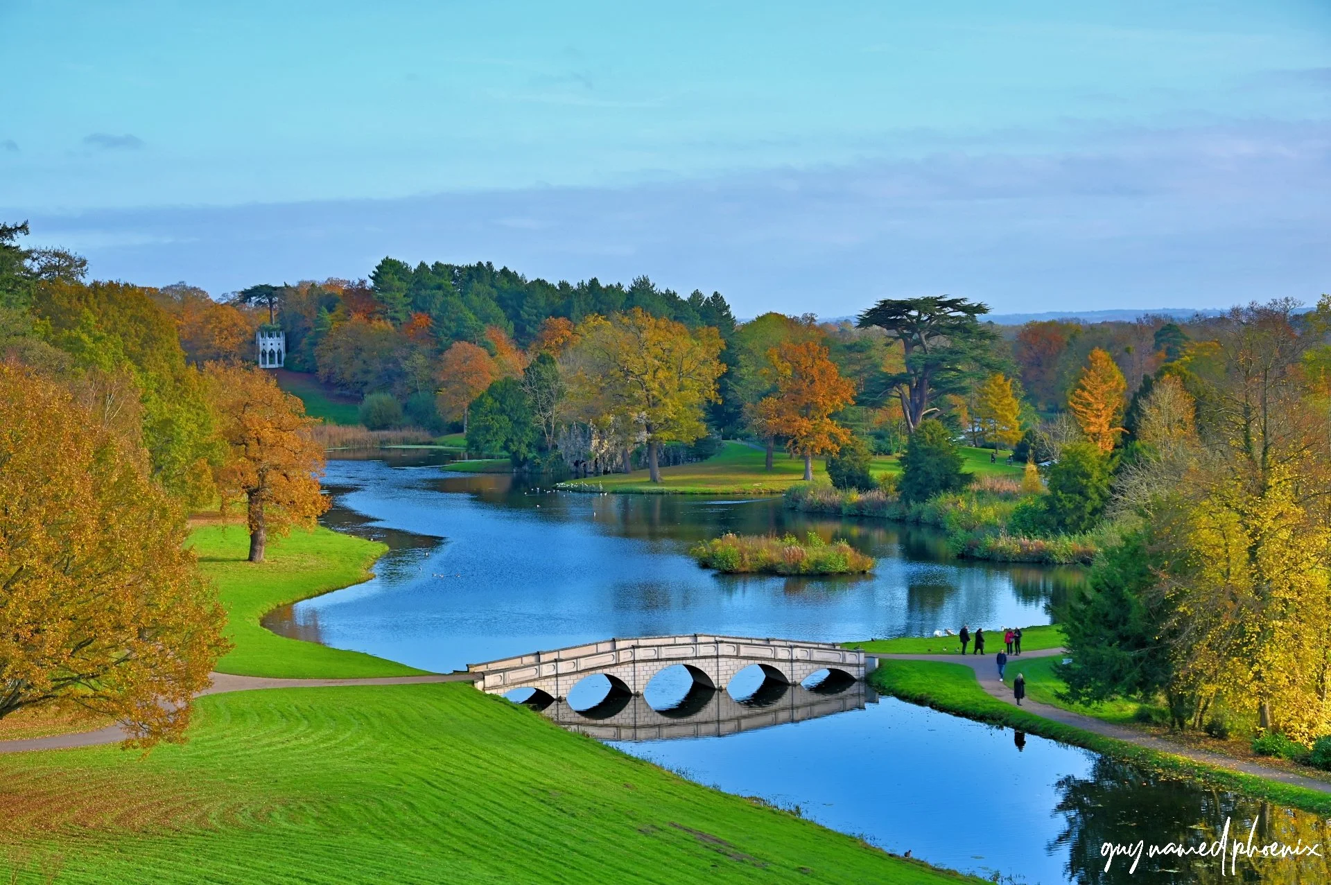 A scenic view of a river running through a park with lush green grass, trees with autumn-colored leaves, and a small white stone bridge. There are walking paths with people, and a distant white building is visible on a hill. The sky is partly cloudy.