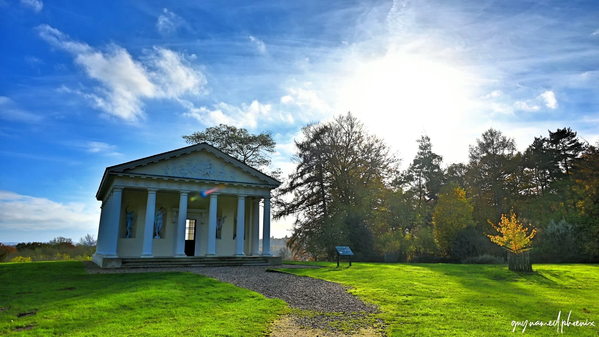 A small Greek-style temple with columns, statues, and a triangular pediment surrounded by green grass and trees, with the sun shining brightly in the sky.