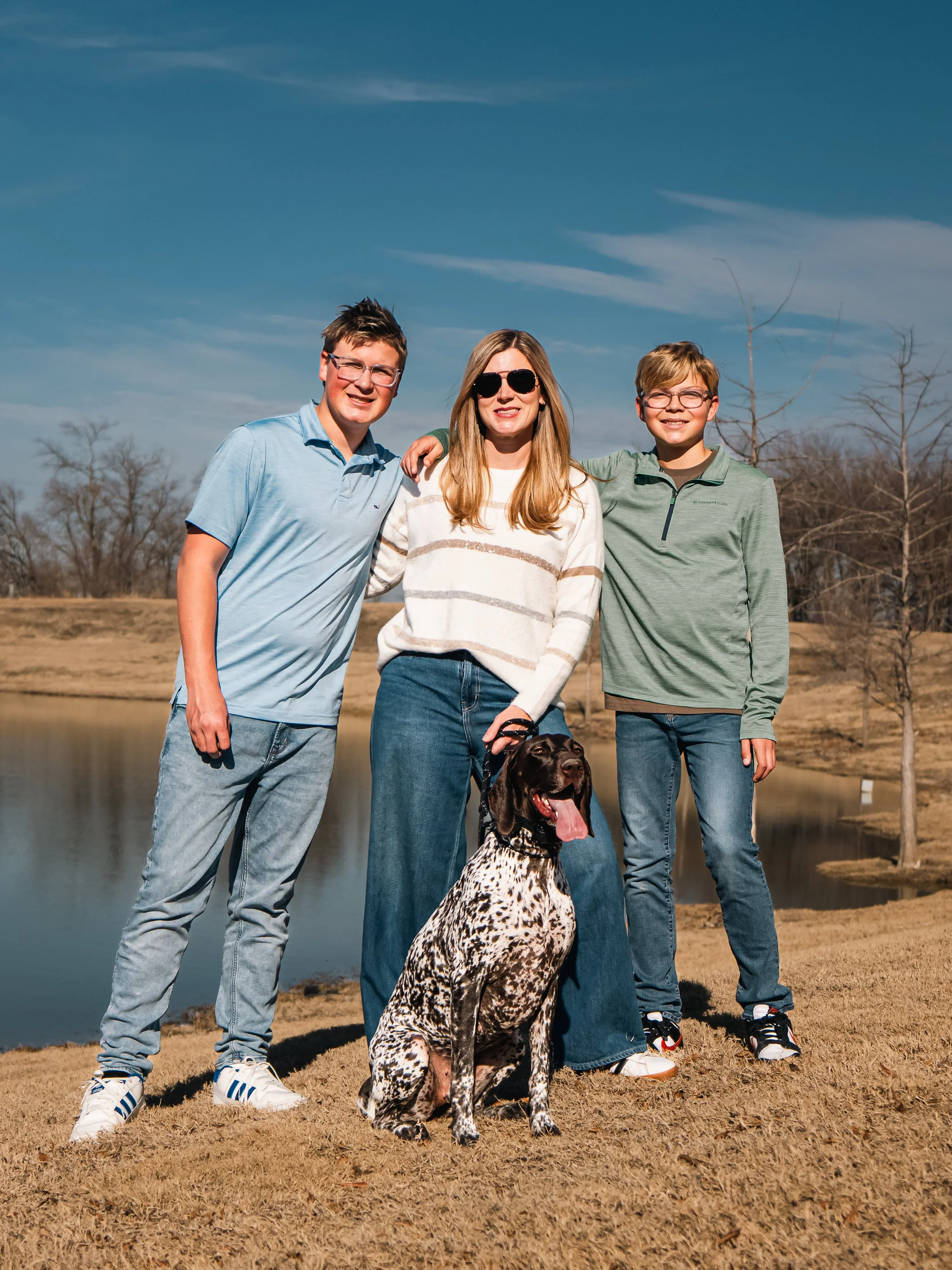 A woman and two boys standing outdoors with a dog by a lake, sunny sky, and leafless trees in the background.