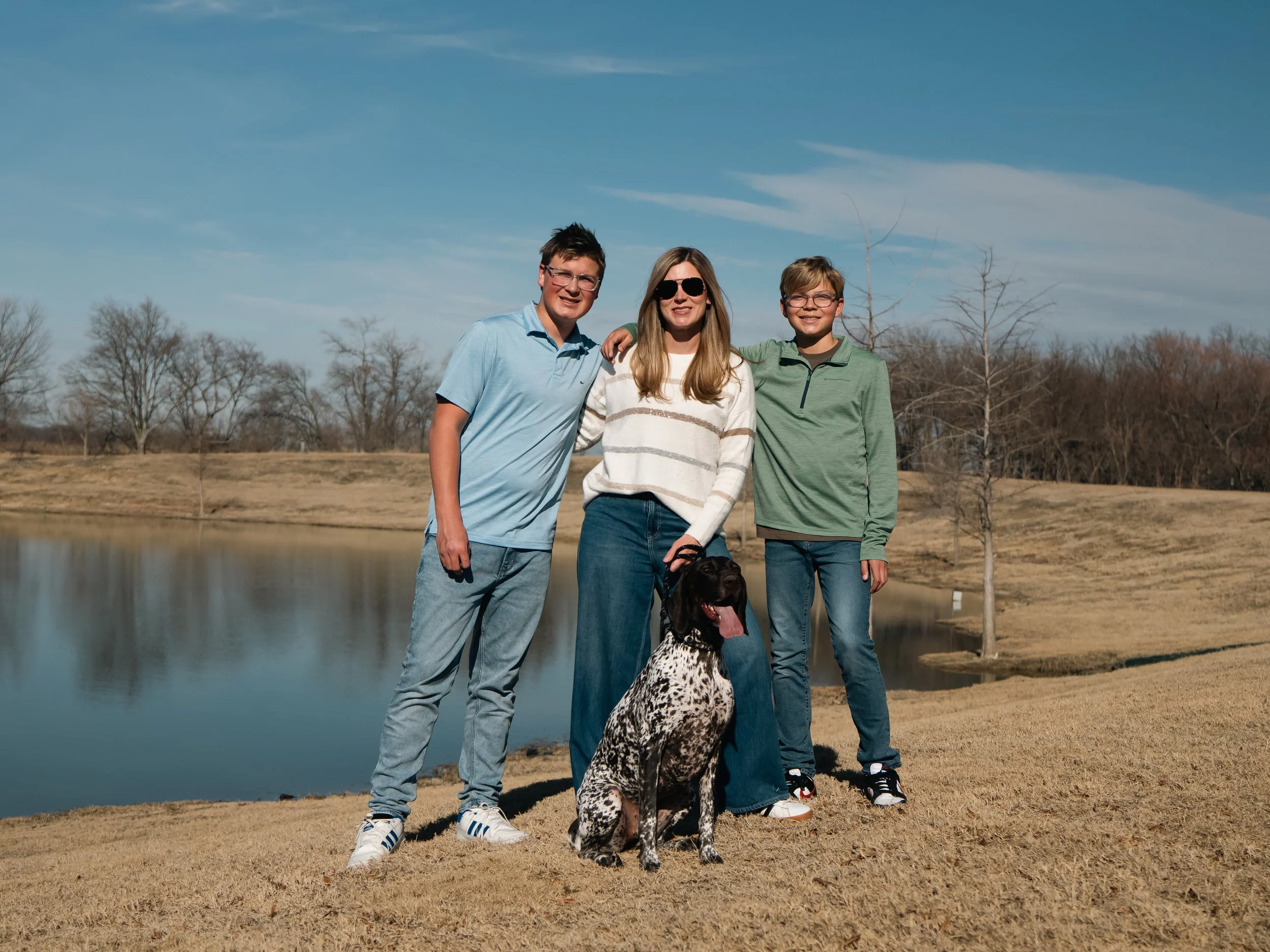 Family of three teenagers and a woman with a dog standing outdoors near a pond, with leafless trees in the background on a clear day.