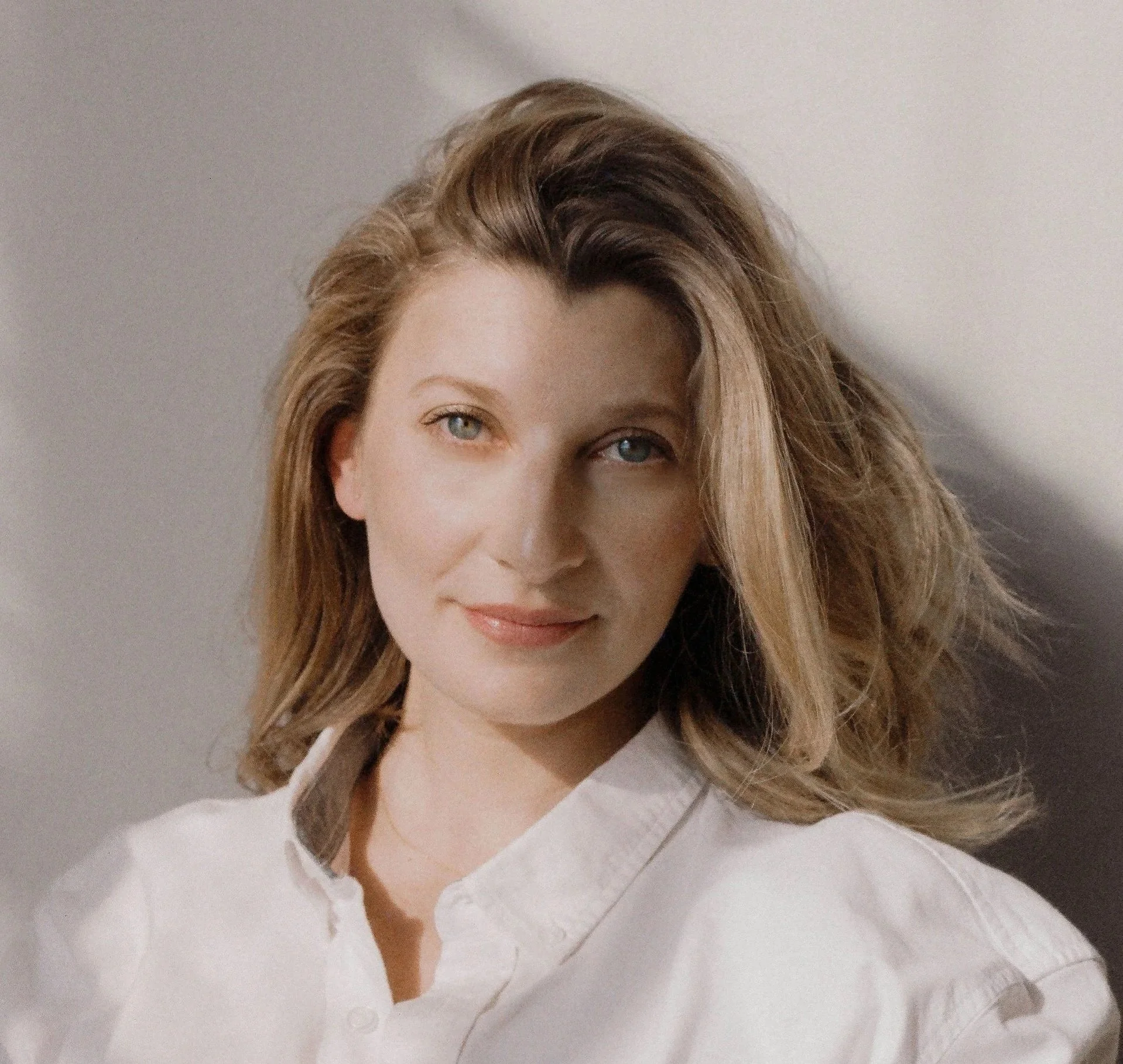 A portrait of a Lisa Krady, woman with wavy, shoulder-length blonde hair, blue eyes, and fair skin, wearing a white collared shirt, smiling subtly. The background is plain and neutral.