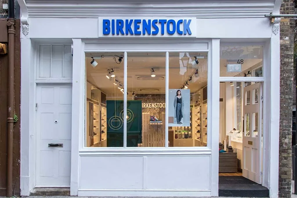 Front view of Birkenstock store with white facade, large glass window displaying shoes and a poster of a woman in blue overalls, and a blue sign with white text reading 'BIRKENSTOCK'.
