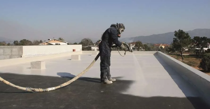 A worker spray-paints a roof with a large hose, wearing protective gear.
