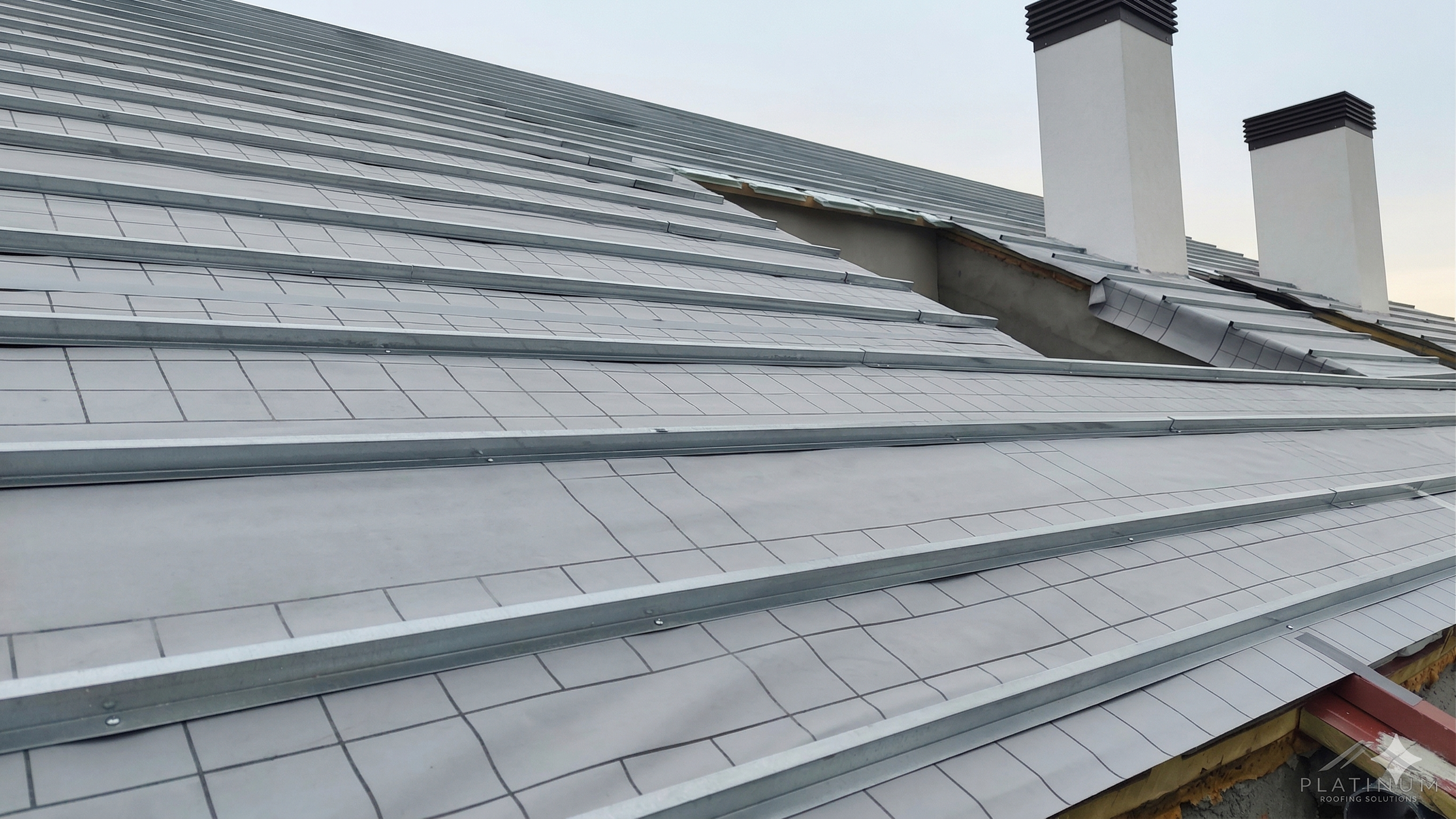 Metal roofing panels installed on a sloped roof with visible black-chimney vents in the background.
