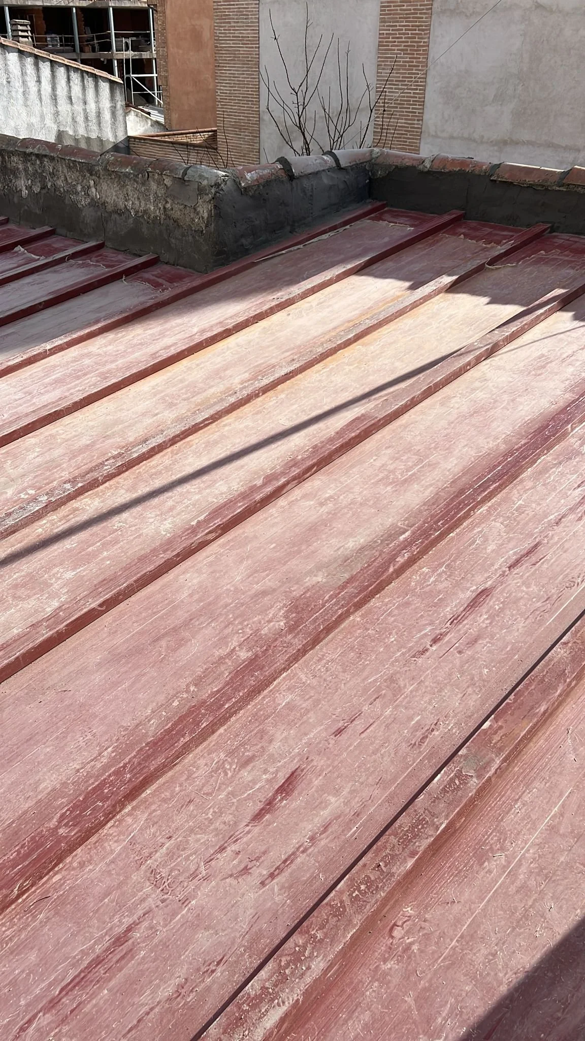 Close-up of a weathered, pink metal roof with raised seams and a brick chimney with some construction materials or scaffolding visible in the background.