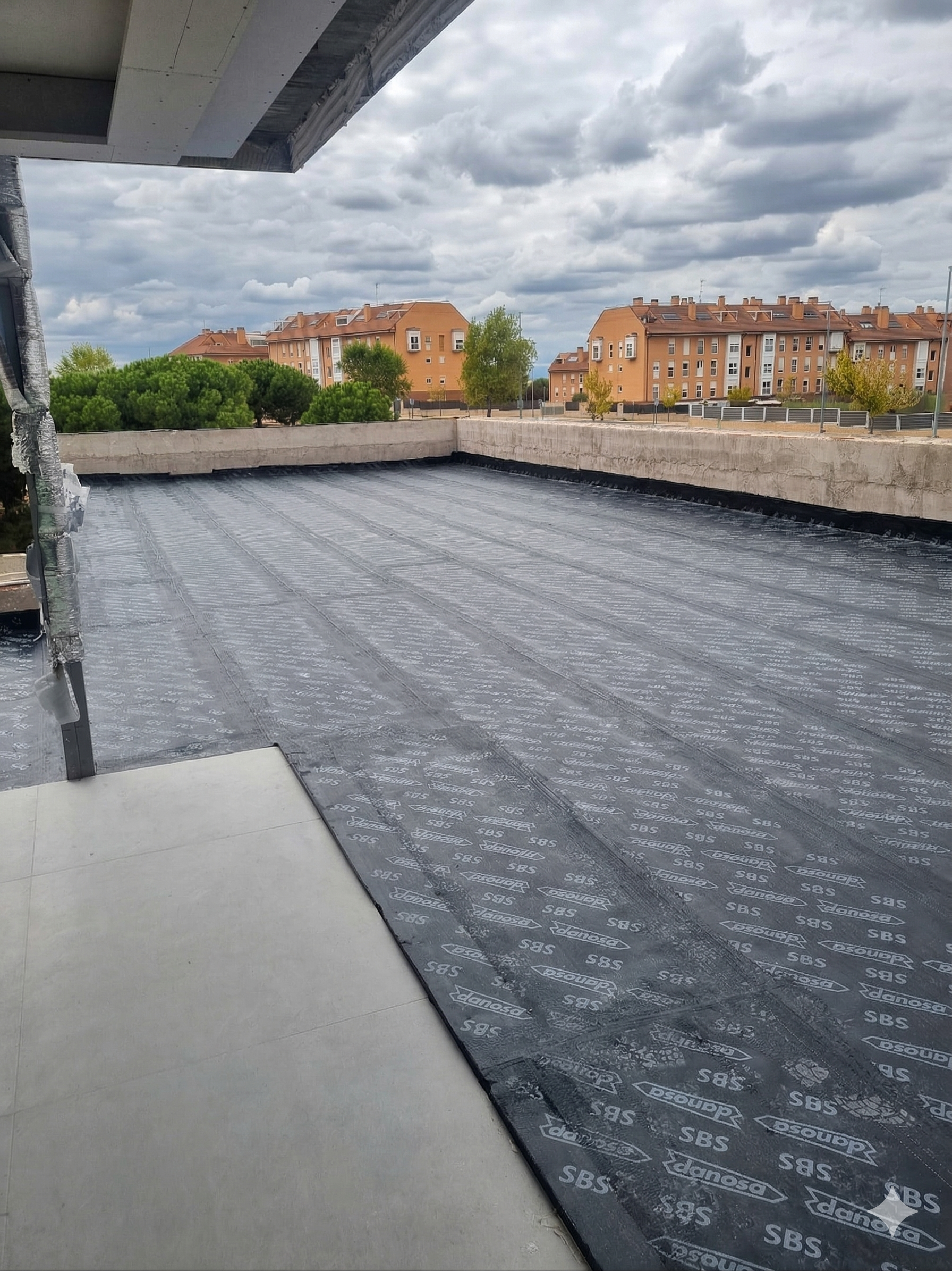 Photo of a building roof under construction with black waterproofing membrane and some residential buildings in the background under a cloudy sky.