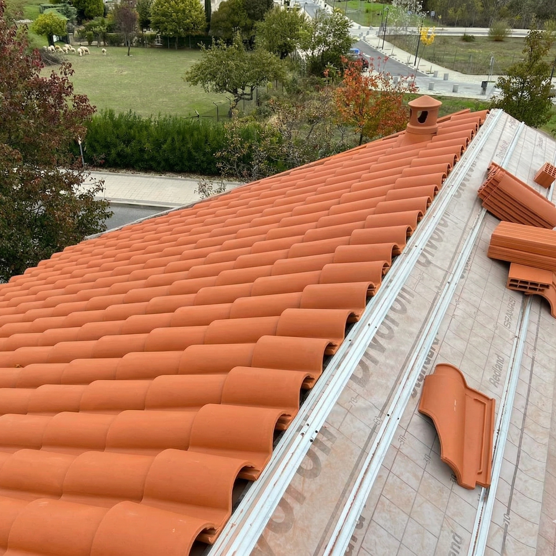 Roof under construction with orange clay tiles, roofing materials, and a chimney, overlooking trees, a park, and a parking lot.