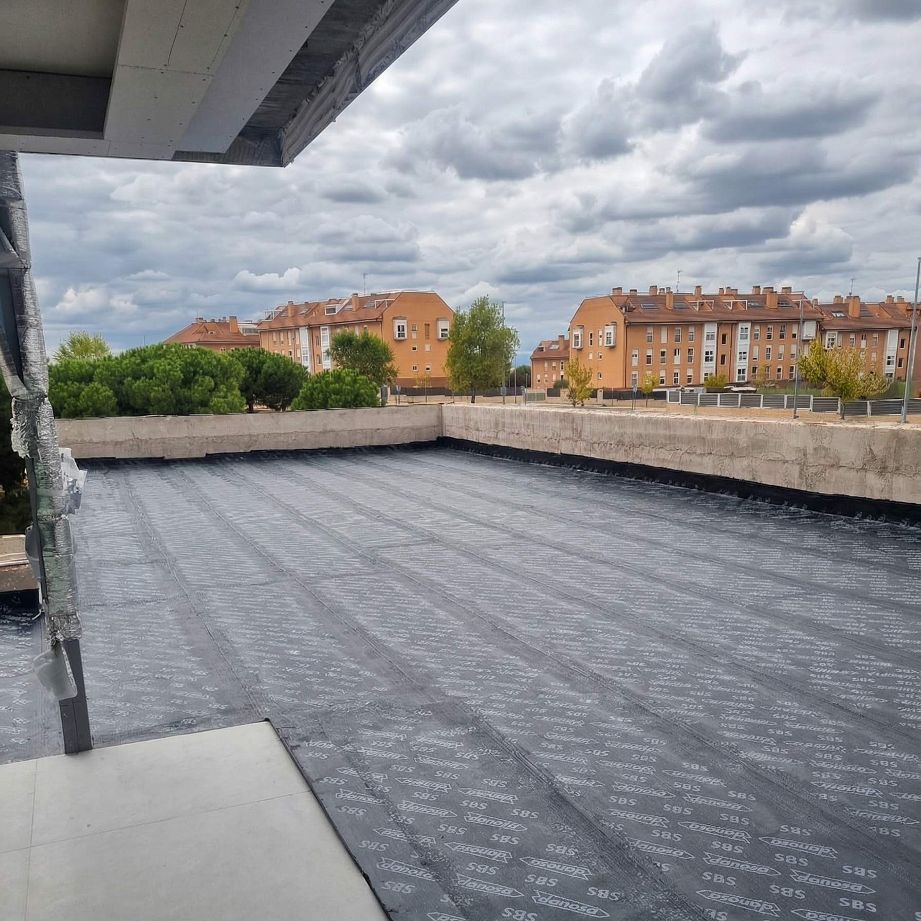 View of a flat rooftop under construction with a black waterproofing membrane, surrounded by a low concrete wall. In the background, there are residential buildings with multiple stories, and green trees under a cloudy sky.