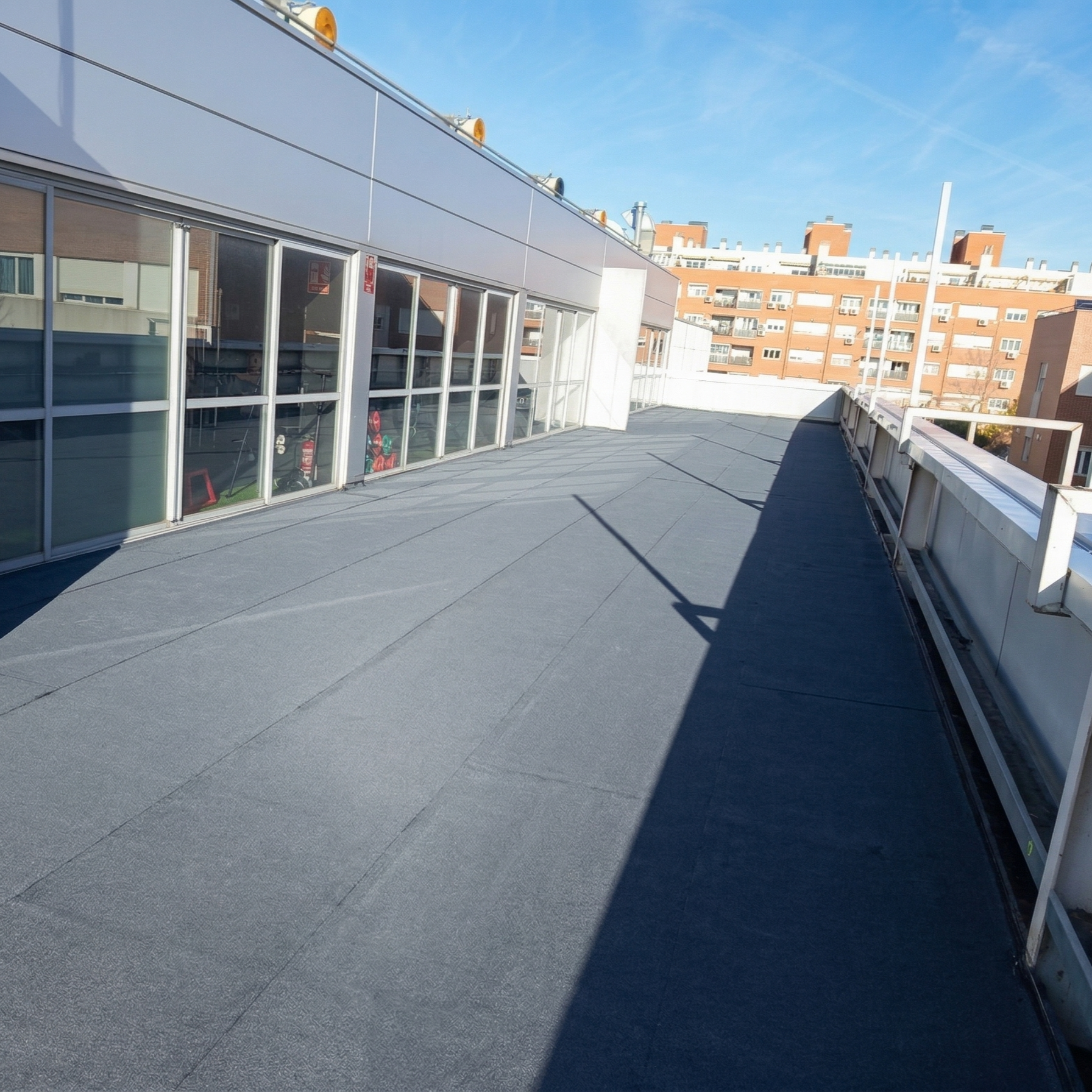 Empty rooftop terrace with glass panels and a clear blue sky, overlooking surrounding apartment buildings.