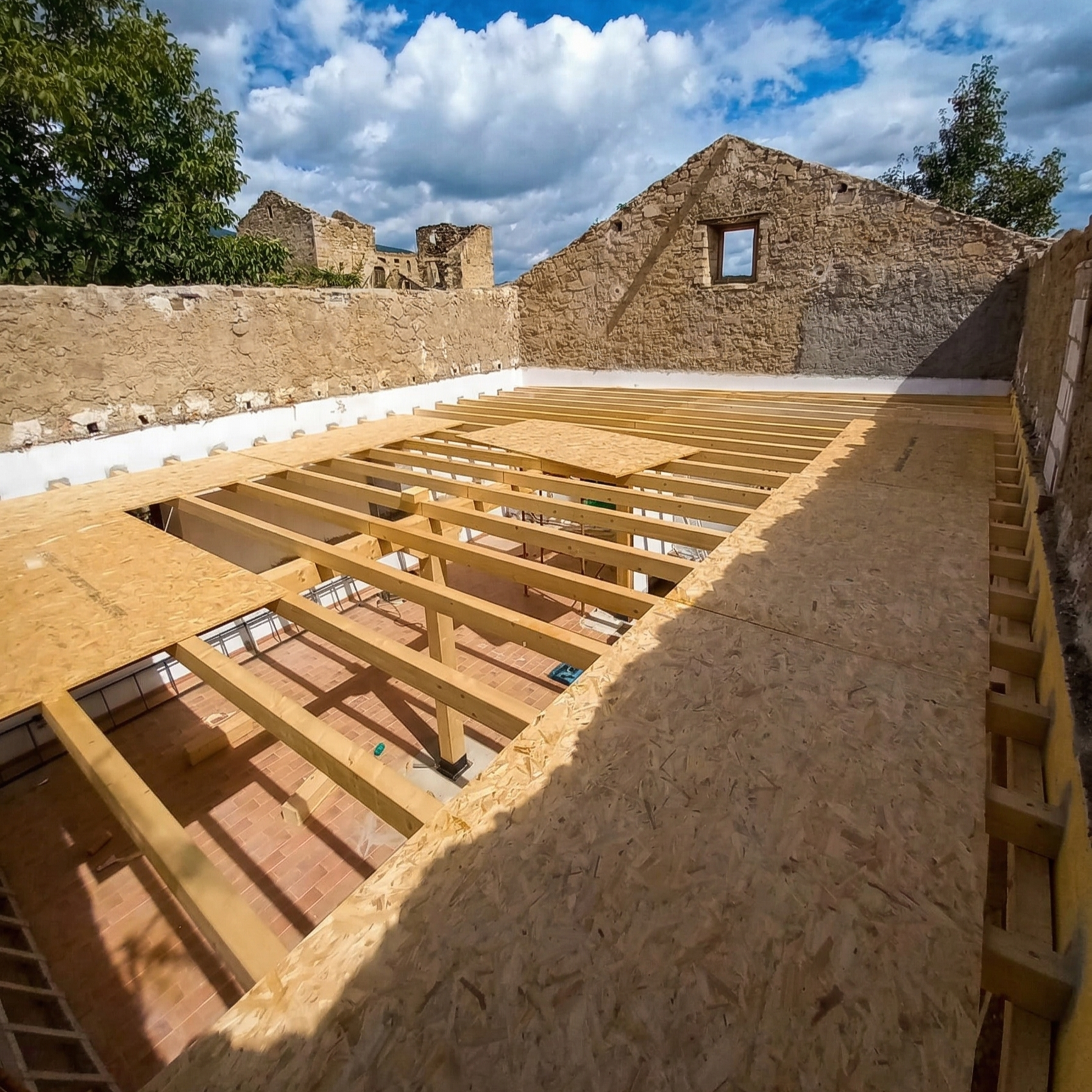 Under construction rooftop with wooden beams and OSB plywood sheathing, surrounded by older stone buildings, under a cloudy sky.